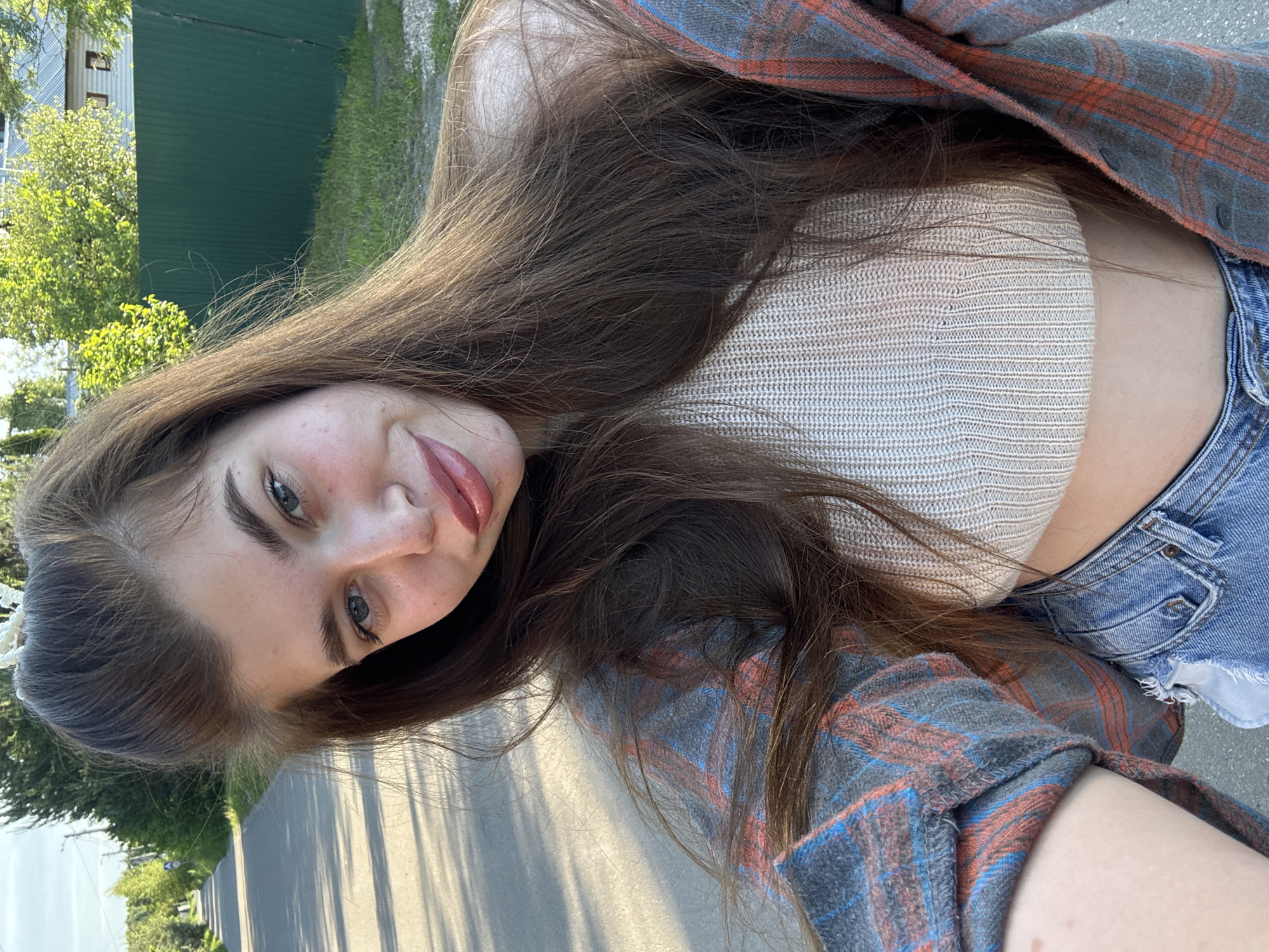 A young woman is captured in a brightly lit outdoor selfie, holding the camera above and slightly to her side, giving a skewed perspective to the background. She has long, dark brown hair flowing around her shoulders and across her chest. Her blue eyes look directly at the camera, and she wears a soft smile with subtly glossed lips. She is dressed in a light-colored, possibly beige or off-white, ribbed knit crop top, layered with an open plaid shirt featuring red, blue, and gray tones. Below the crop top, a small portion of blue denim shorts is visible.
The background shows a clear, sunny day with green foliage, including trees and bushes, suggesting a lush outdoor environment. A dark green building or fence is partially visible behind her. A paved road stretches into the distance, marked with distinct, elongated shadows, indicating it is likely morning or late afternoon. The overall scene conveys a casual, relaxed atmosphere in what appears to be a suburban or rural setting, specifically Мещёры, Russia. There is no visible text in the image.