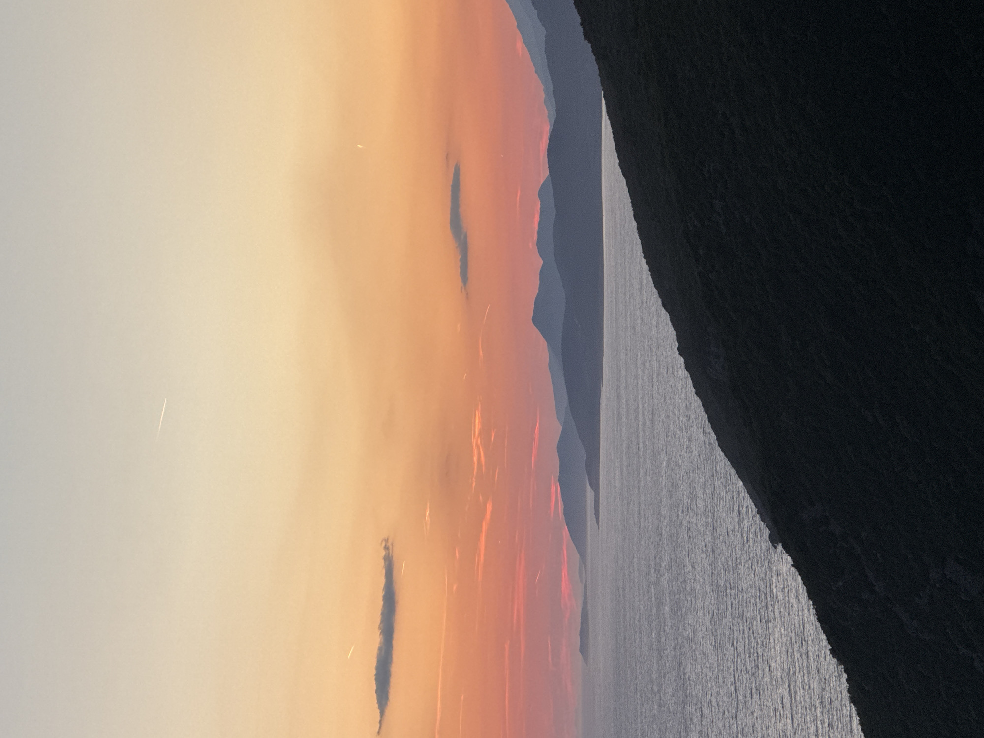 This is a photograph of a sunset over the Adriatic Sea, likely taken from a vantage point overlooking the coastline of Zagora, Montenegro. The sky is a gradient of colors, transitioning from a pale yellow at the top to a vibrant orange and pink near the horizon. Several wispy clouds are scattered across the sky, some tinged with the sunset's hues. In the distance, a series of silhouetted mountain ranges stretch across the frame, their forms softened by the atmospheric haze. The sea in the foreground is a muted gray-blue, reflecting the subdued light of the fading day. A dark, densely vegetated hillside slopes down from the lower right corner of the frame, meeting the water's edge. There are no people or man-made structures immediately visible. The scene evokes a sense of tranquility and natural beauty.