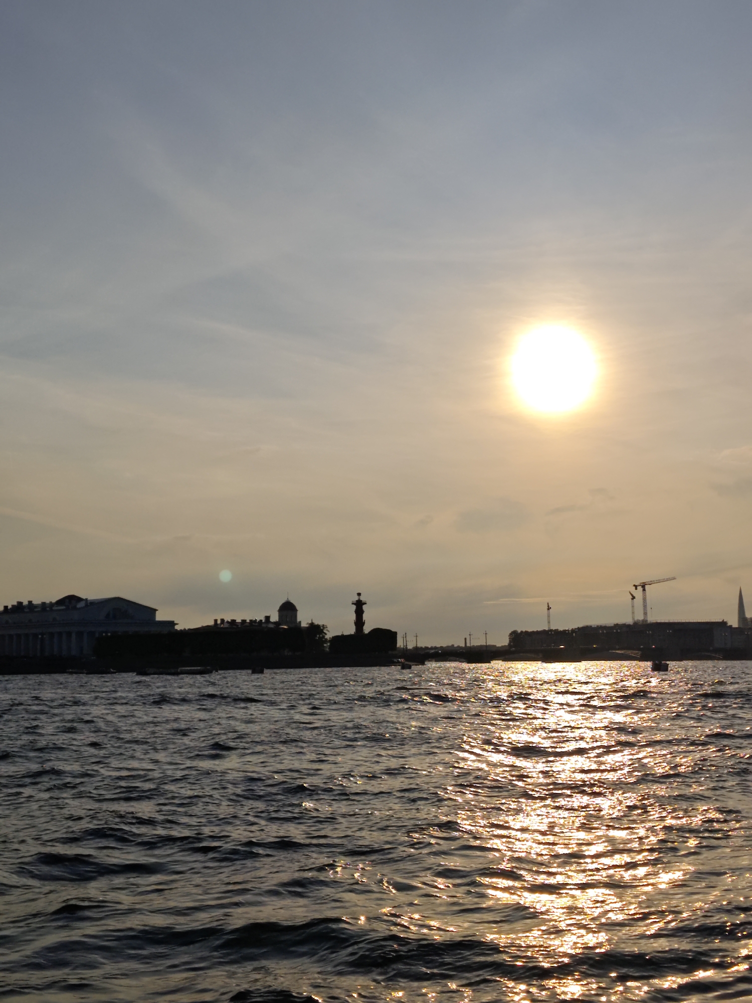 The image depicts a sunset over the Neva River in Saint Petersburg, Russia. The sun, a bright disc, is positioned in the upper right quadrant of the sky, casting a golden glow on the water's surface. Silhouetted against the sky are various buildings and structures on the far bank. Prominent among these are a long, colonnaded building on the left, a domed structure, and a tall rostral column. In the distance, cranes suggest ongoing construction. The water is choppy, reflecting the sunlight in shimmering patterns. Several small boats are visible on the water. The overall scene conveys a tranquil, late afternoon or early evening atmosphere.