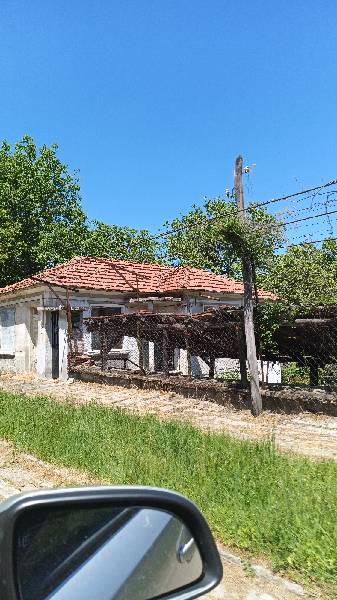 The image depicts an aged, single-story structure with a red tiled roof, located in Gornoslav, Bulgaria. The building appears to be in a state of disrepair, with a chain-link fence partially obscuring the front facade. The fence is supported by wooden posts, one of which is also a utility pole with electrical wires. Green foliage and trees surround the building. In the foreground, tall grass is visible, and the side mirror of a vehicle is in the lower left corner. The sky is a clear blue. The setting appears to be rural.
