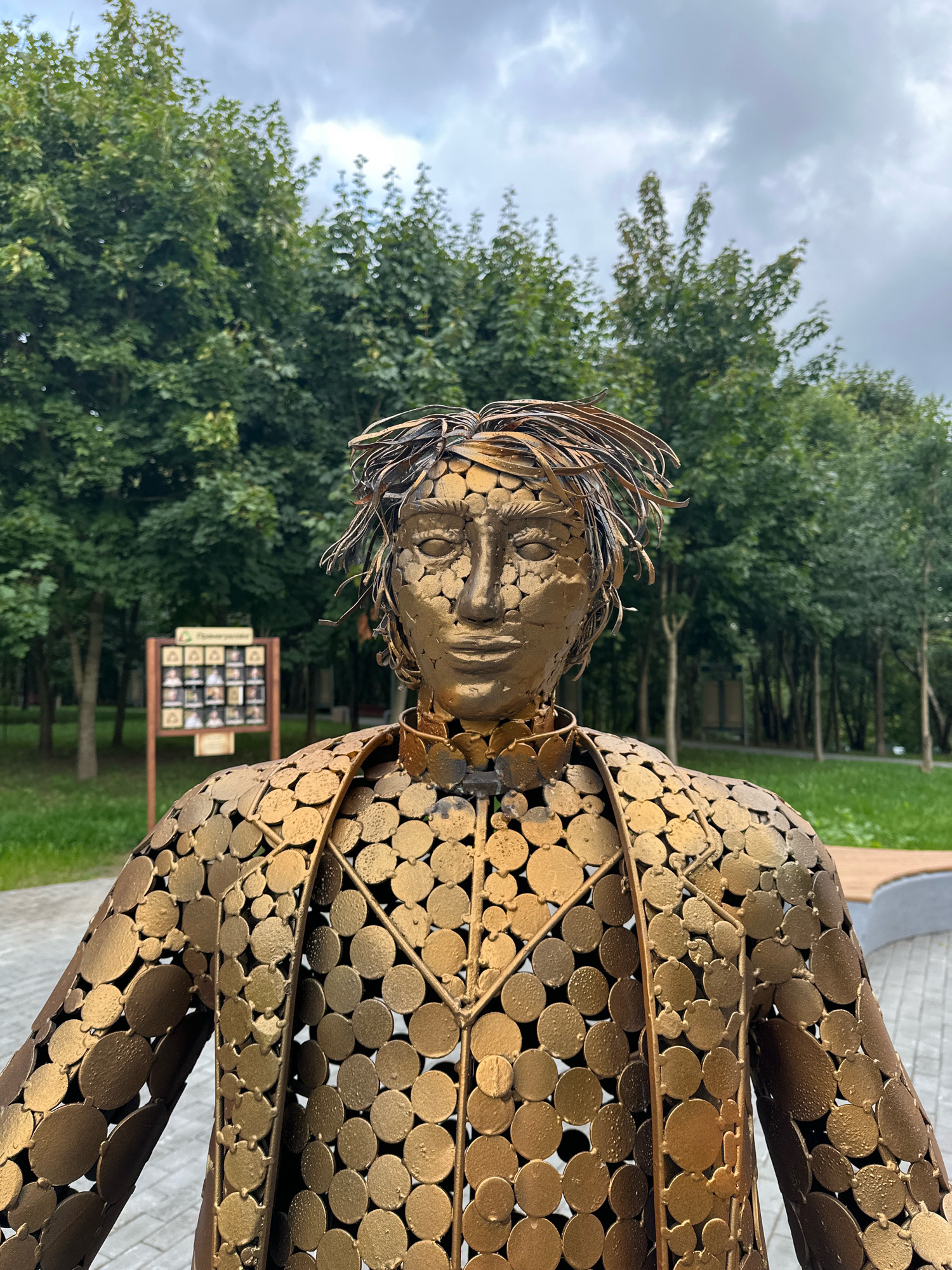 The image shows a close-up of a large, golden, metallic sculpture of a person's torso and head, which is located in the city of Minsk, Belarus. The sculpture is made of many small, circular metal pieces that form the clothing and body of the figure, with strands of metal wire creating the hair. The face of the sculpture is detailed, with sculpted eyes, nose, and mouth. Behind the sculpture, a park setting is visible with green trees and a cloudy sky. A wooden display board with several framed photographs is also present in the background, to the left of the sculpture. The ground in the foreground appears to be paved.