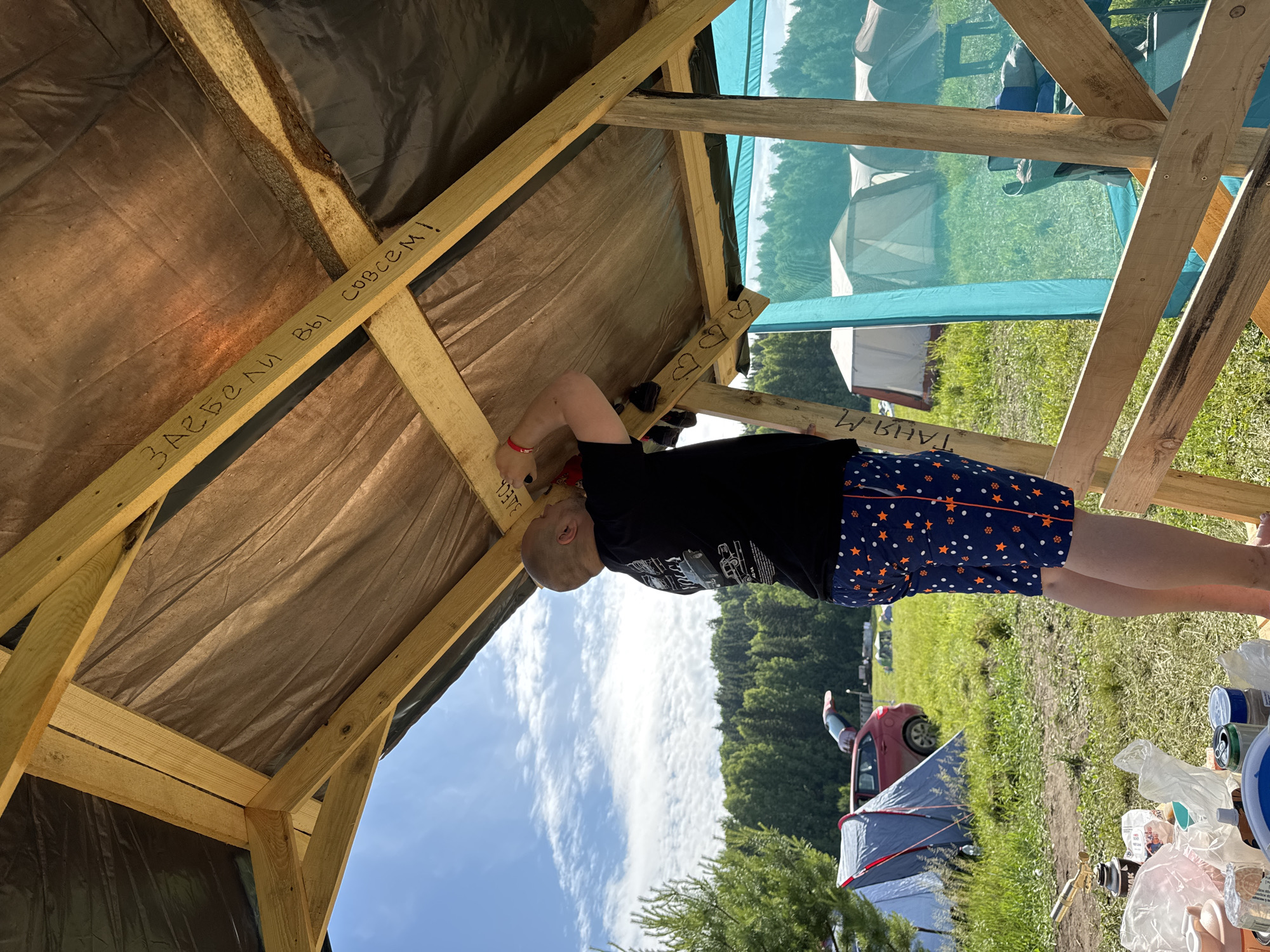 The image depicts an outdoor scene in Большая Кивара, Russia, featuring a male individual working on a wooden shelter under a clear sky with scattered clouds. The man, seen from the waist up, is bald and wearing a black t-shirt with white graphic prints on the back and dark blue shorts with orange stars and white polka dots. He is standing on a wooden frame, reaching upwards with both hands to fasten material to the underside of a roof-like structure. A red wristband is visible on his left wrist.
The structure he is working on consists of rough-hewn wooden beams and planks, forming a simple roof frame. The underside of the roof is covered with a dark, tarp-like material, with a lighter brown fabric layer visible beneath it in some sections. Text written in black marker is present on the wooden beams: "ЗАЕБАЛИ ВЫ СОВСЕМ!" is visible on an upper horizontal beam, and "ТАНЯ М" is partially visible on a vertical post to the left. Three hand-drawn heart shapes are also marked on a vertical post below the man's left arm.
In the background, a grassy field extends to a distant treeline of dense green forest. Several tents are pitched across the field, varying in size and color, predominantly light gray or white. A red car with its trunk open is parked behind one of the tents on the left. To the right of the man, a turquoise net or fabric wall of another structure is visible. In the bottom left foreground, a collection of items, including plastic bags, a metal can, a plastic container with a blue lid, and a small portable gas stove, are arranged on what appears to be a flat surface or pallet on the grass. The overall lighting suggests a bright, sunny daytime environment.