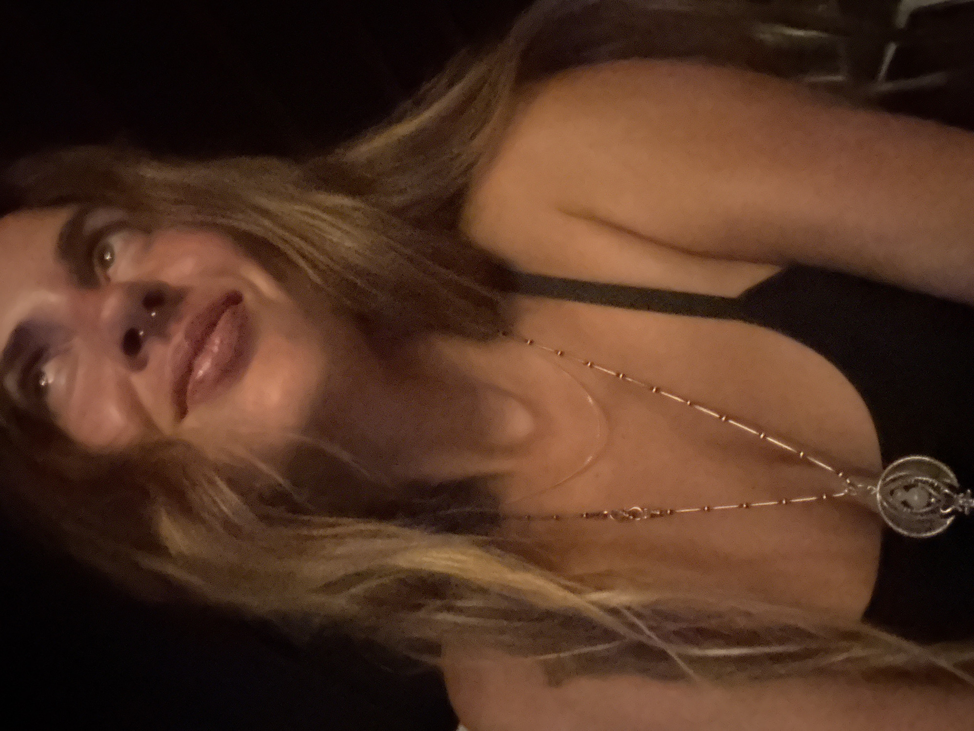 A woman with blonde hair and a nose ring looks up and to her right, with a slight smile. She is wearing a black tank top and a long necklace with a circular pendant. The background is dark and out of focus, suggesting an indoor or nighttime setting. There are no visible landmarks or environmental clues to indicate a specific city or location.