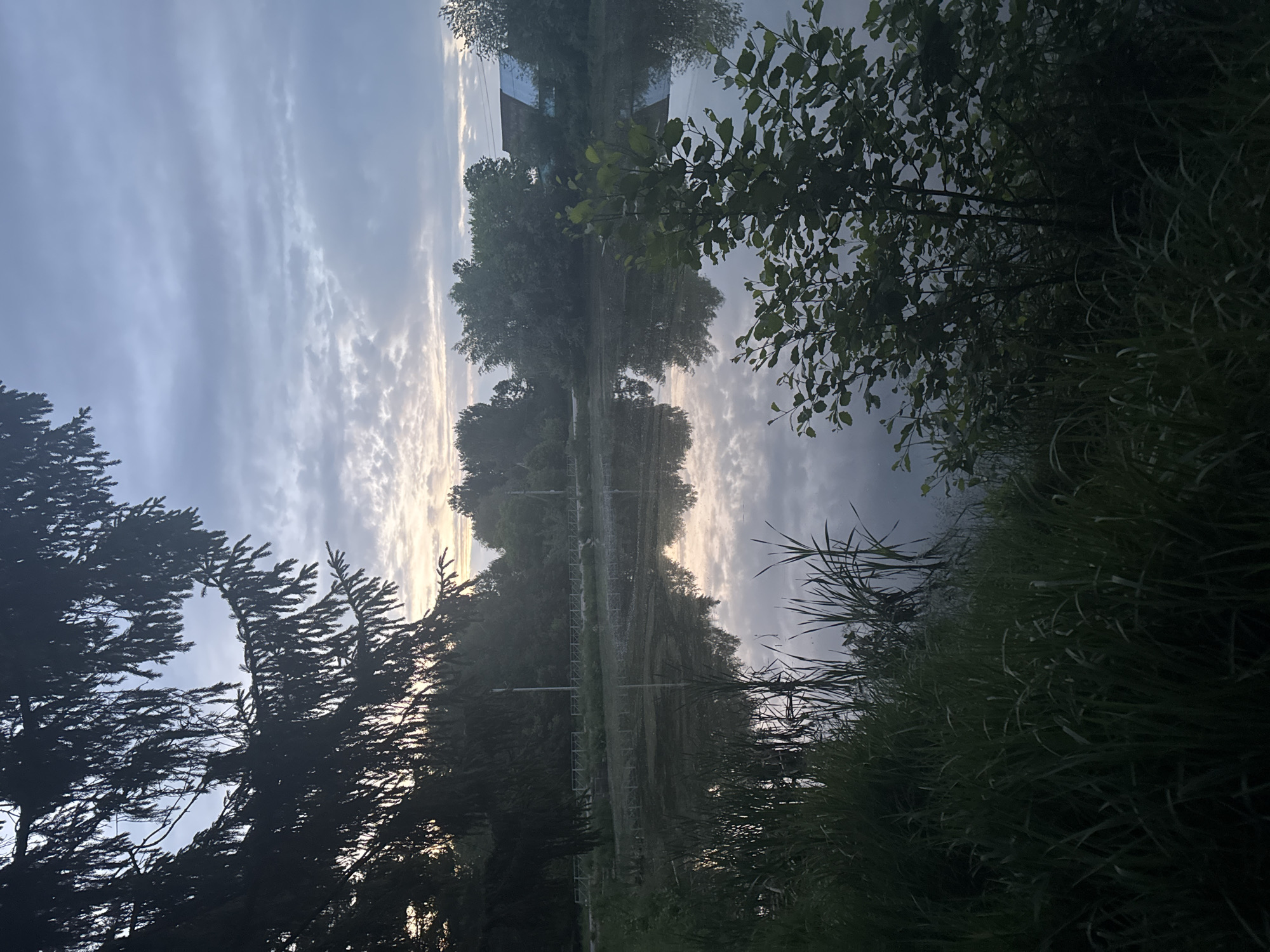 The image shows a body of water reflecting a sky with layered clouds and a hint of sunlight. A line of trees and vegetation is visible along both edges of the water. An architectural structure, likely a building, is reflected in the water. A fence is visible in the background of the shot. The photograph was taken near the city of Chaikovsky, Russia.