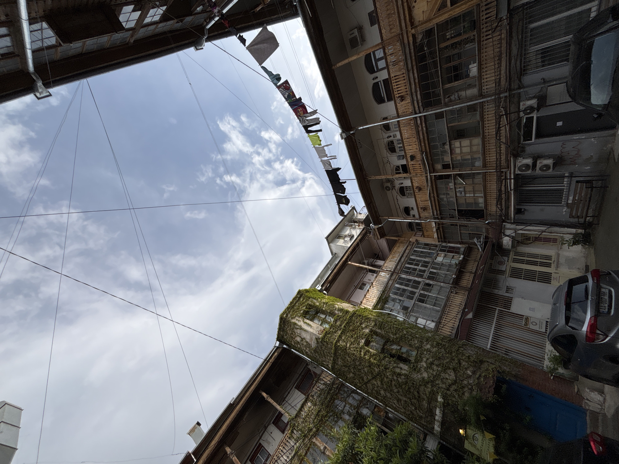 An upward-looking view captures the narrow sky between old, multi-story buildings in what appears to be an urban alley or courtyard. The architecture features aged stone or stucco walls, wooden balconies, and numerous windows, some of which are grimy or fitted with metal grates. One prominent building on the left is heavily adorned with climbing ivy.
A clothesline laden with various garments, including t-shirts and shorts, stretches across the sky, indicating laundry drying. Numerous utility wires and cables crisscross the bright, partially cloudy sky. Air conditioning units and drainage pipes are visible on the building facades.
On the right, a silver car is parked, with its rear visible, bearing a Georgian license plate "AA-007-HH", confirming the location as Tbilisi, Georgia. A hint of a red rear light from another vehicle is visible further down. At the bottom, green foliage from trees or bushes is present, and a yellow sign with partially legible text, possibly "VINTAGE", hangs near the bottom right corner. The scene is brightly lit, suggesting daytime, under an overcast sky with scattered clouds. The overall impression is one of an authentic, lived-in urban environment.