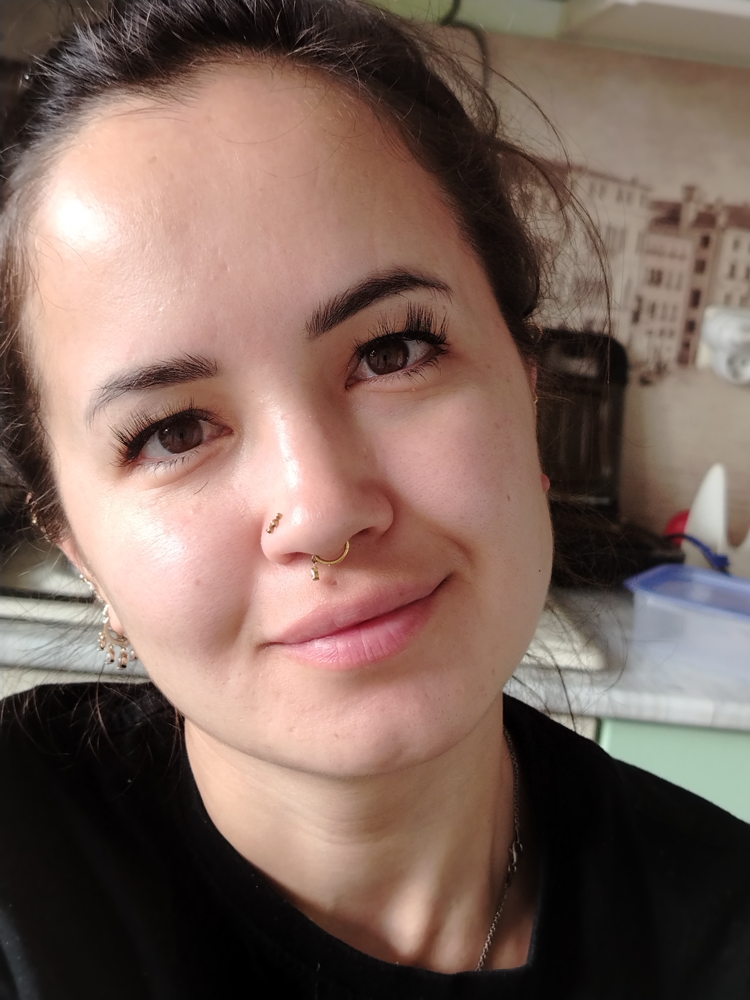 A close-up, eye-level, medium shot shows a young woman with dark hair, dark eyes, and fair skin. She is looking towards the camera with a slight smile. Her hair is pulled back, and she has on false eyelashes and two nose piercings. She is wearing a black shirt. The background appears to be a kitchen, with cabinets and a decorative backsplash visible. There is a blue plastic container on a counter in the background. The image was taken indoors, possibly in Noviy, Russia.