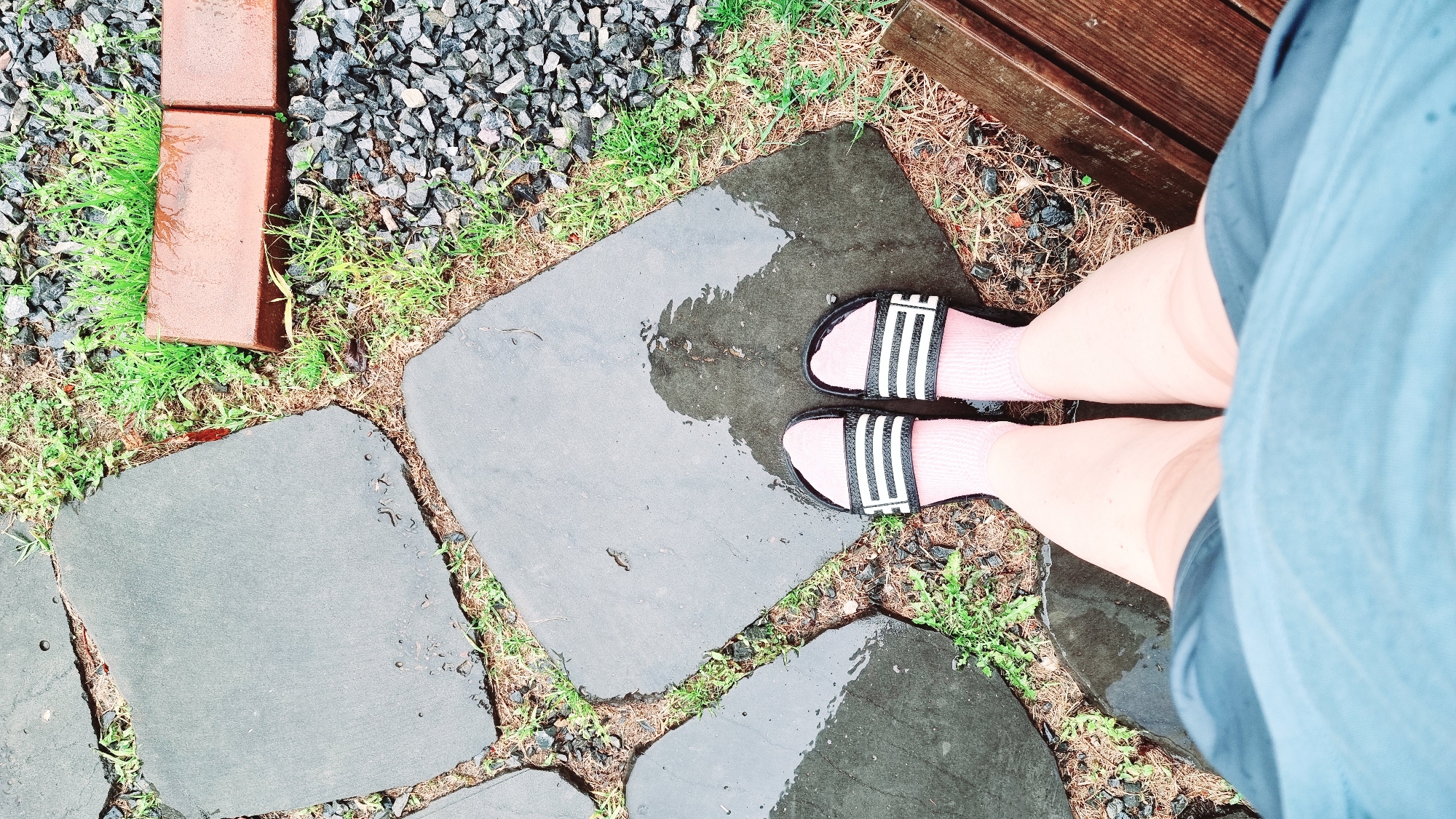 The image shows a first-person perspective of someone standing on a wet stone pathway in Dunnae-myeon, South Korea. The pathway is made of irregular gray stone slabs with grass and weeds growing in the gaps between the stones. The person is wearing pink socks and black and white striped sandals. A small section of a wooden bench is visible on the upper right side of the image. To the left of the stone pathway is an area covered with gray gravel and green grass, with a few red bricks near the edge. The person's legs and a portion of their light blue shorts are visible in the right side of the frame. The environment appears to be wet, possibly after a rain.
