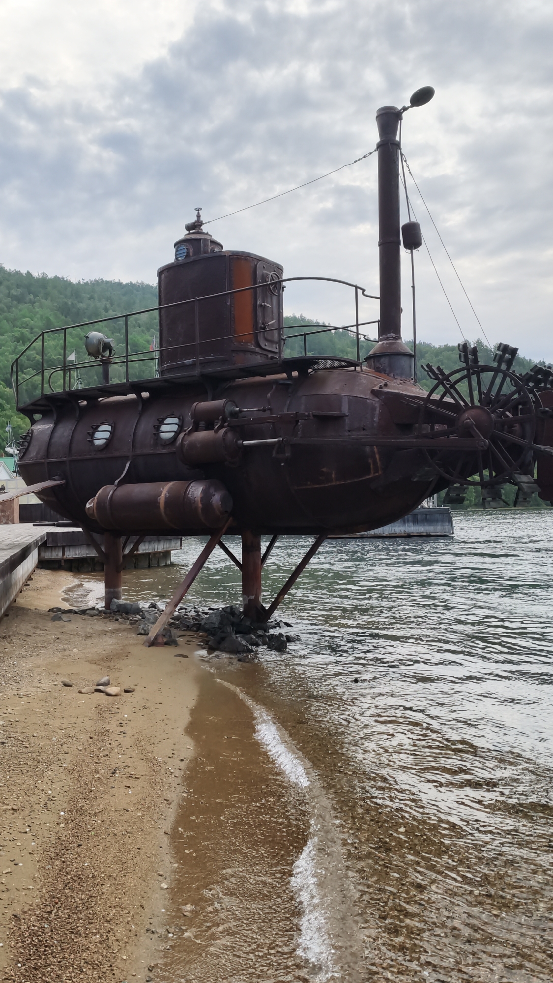 An abandoned, rusty, steampunk-style submarine sits on the sandy shore next to a body of water in Nikola, Russia. The submarine is elevated by several metal supports and appears to be made of riveted metal. It has multiple portholes, a large smokestack, and what appears to be a paddle wheel at the stern. A wooden pier extends from the left side of the frame, leading towards the submarine. The water is calm with small waves lapping at the shore, and the background features a tree-covered hillside under a cloudy sky. The overall scene depicts a static, weathered structure in a natural, waterfront setting.