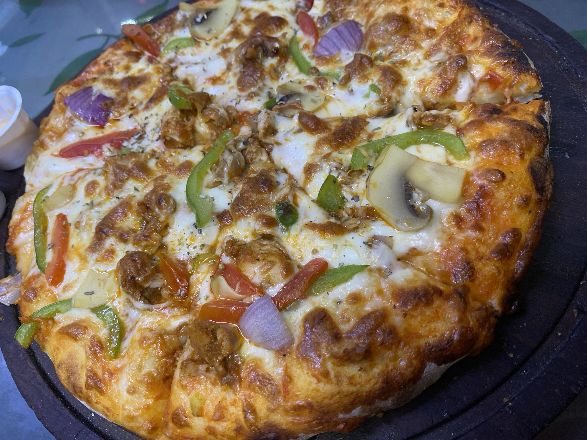 A close-up, high-angle shot depicts a pizza on a dark wooden platter. The pizza is topped with melted mozzarella cheese, browned ground meat, sliced mushrooms, green and red bell peppers, and red onions. The crust is golden brown. A small, white container with a creamy substance sits near the left edge of the platter. The photo was likely taken in Dhaka, Bangladesh. No people are visible.