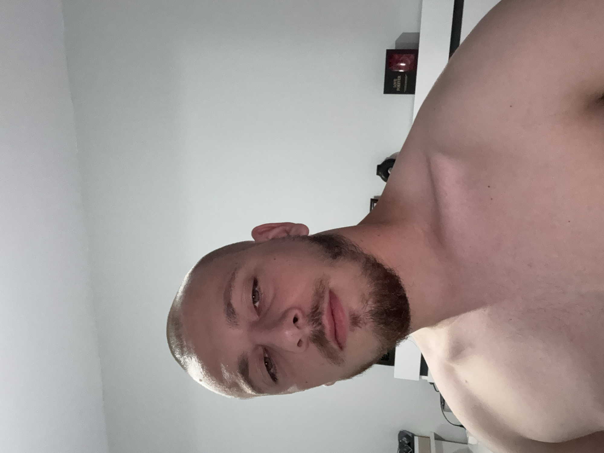 The image depicts a close-up, chest-up view of a young adult male with a shaved head and a short beard. He is looking directly at the camera with a neutral expression. His skin is fair, and his shoulders and upper chest are visible, appearing bare. The background is a plain, light-colored wall. On a shelf or dresser behind him, to the right, is a small, dark-colored box with the words "LOVE FOREVER" printed on it, along with a red object visible through a transparent window. To the left, some indistinct objects are partially visible, including what appears to be shelving. The lighting suggests an indoor setting, likely a bedroom. There is no visible indication of the location being Stara Zagora, Bulgaria.