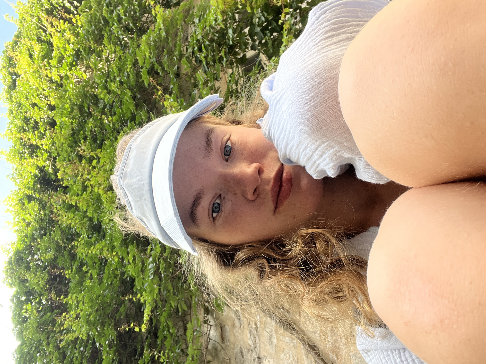 The image is a low-angle selfie of a fair-skinned person with light blonde, curly hair, blue eyes, and freckles, wearing a light blue visor and a light blue, textured top. The person's legs are visible in the upper right corner of the frame. In the background, there is greenery consisting of dense ivy-covered stone, and an unclear stone structure. Given the context of Budva, Montenegro, it is likely that the greenery is related to the city's coastal environment, possibly along the Old Town walls or in one of the parks along the coast. The subject is looking directly at the camera.
