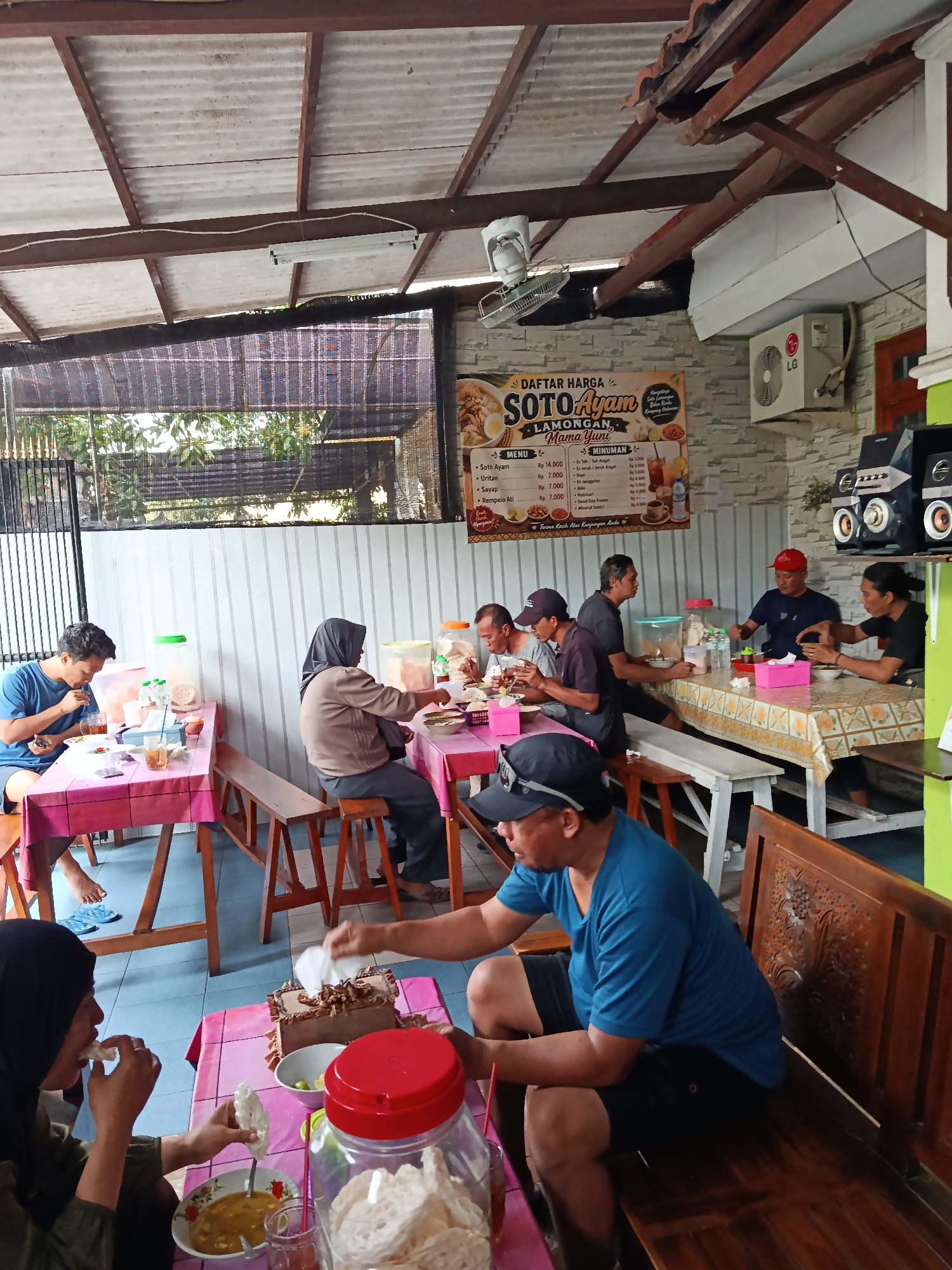 This image depicts an outdoor dining area of a restaurant in Keboansikep, Indonesia. Several patrons are seated at tables, enjoying meals and beverages. In the foreground, a man wearing a blue t-shirt and black shorts sits on a wooden bench. To his left, a woman in a hijab is eating, holding a piece of crackers. A table between them is set with a bowl of soup, glasses, and a container of crackers.
Further into the scene, other diners are visible. A man in a blue t-shirt is seated at a table covered with a pink tablecloth, eating and holding a phone. To his right, a woman in a brown jacket and hijab is seated. A group of men are seated at a longer table in the background, engaged in conversation and eating.
The restaurant appears to have a simple, open-air setup with a corrugated metal roof supported by wooden beams. A menu board displaying "DAFTAR HARGA SOTO AYAM LAMONGAN Mama Yuni" is prominently displayed on the wall, along with images of food and drinks. An air conditioning unit is mounted on the wall, and a fan is visible near the ceiling. The overall environment suggests a casual dining establishment.