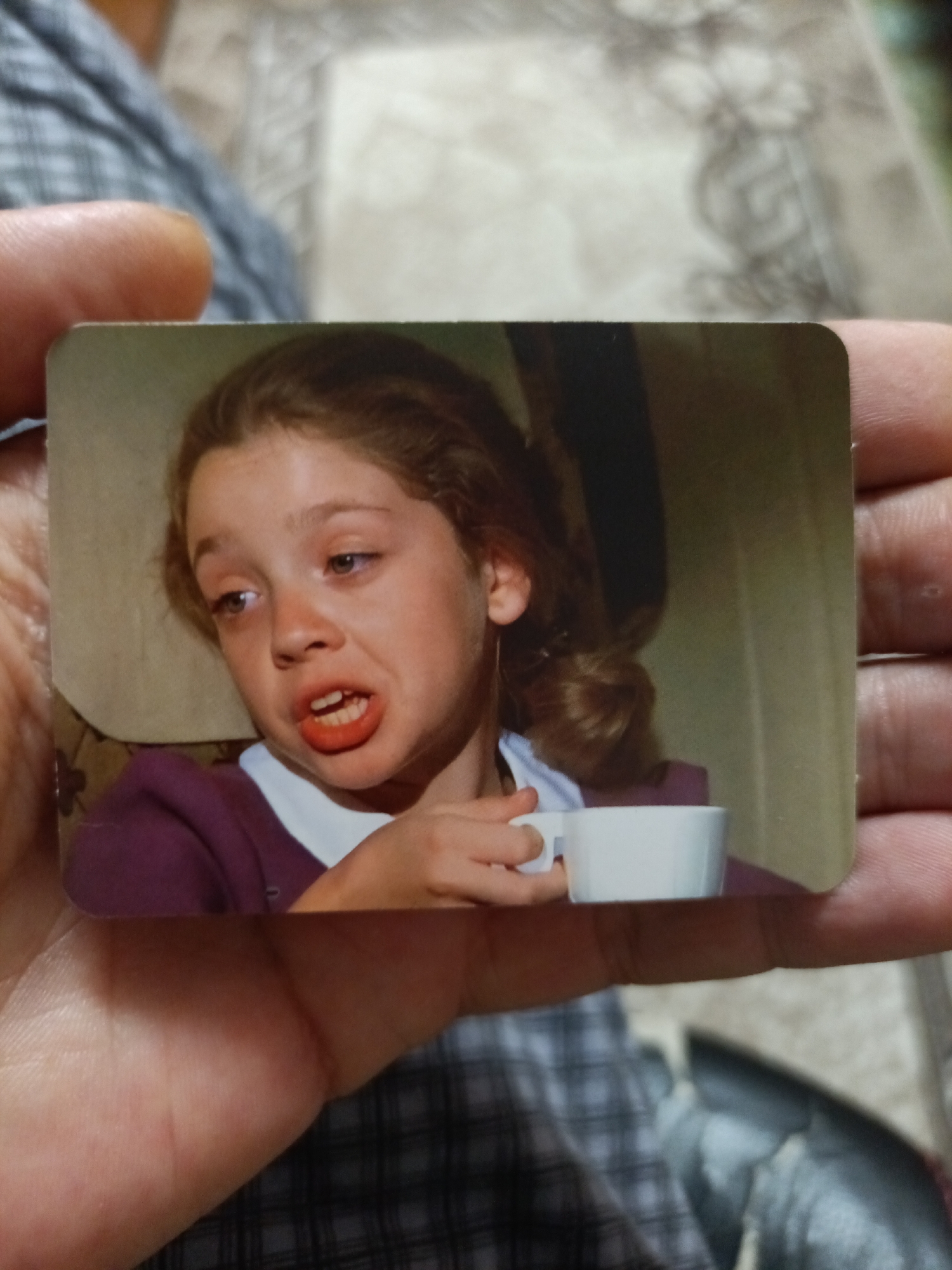 A close-up photograph shows a person's hands holding a rectangular, glossy photograph of a young girl with auburn hair, fair skin, and a distressed expression. The girl is wearing a purple top with a white collar and is holding a small white cup. Her mouth is open, revealing her teeth, and her eyes are wide. The background of the photograph is blurred and appears to be an interior setting. The hands holding the photograph are of a person with light skin. The photograph is positioned over a dark checkered fabric.
