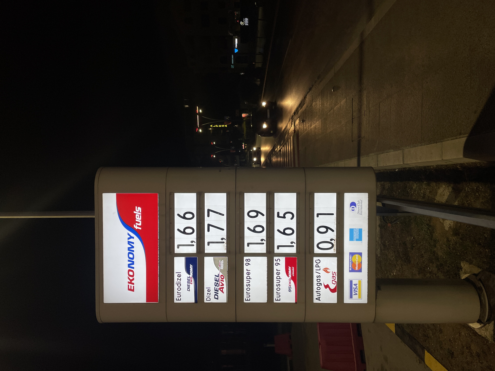 This nighttime image captures an illuminated digital price display for an "EKONOMY fuels" gas station, located in the city of Unknown, Unknown. The vertical sign, primarily white with red and blue accents, displays fuel types and prices: "Eurodizel" (Diesel EKONOMY) at 1.66, "Dizel" (Diesel AVIO Double Filtered) at 1.77, "Eurosuper 98" at 1.69, "Eurosuper 95" (95 EKONOMY unleaded) at 1.65, and "Autogas/LPG" (E-gas) at 0.91. Below the fuel prices, logos for accepted payment cards are displayed: VISA, Eurocard/Mastercard, American Express, and Diners Club International. The base of the sign stands on sparse groundcover with a yellow-painted line visible to the left.
To the right of the price sign, a paved road extends into the background, with its surface reflecting light, indicative of wet conditions. Two vehicles with illuminated headlights are traveling away from the viewer on the road. A white-striped pedestrian crosswalk is visible across the road in the mid-ground. Along the right edge of the frame, a concrete sidewalk borders the road. In the distance, streetlights illuminate the scene, and a vertical sign with yellow illumination displays the text "ALPESA". Further down the street, a blue-lit sign, possibly displaying "PLAZA," is partially visible, along with other distant lights and structures. On the far left, behind the price sign, red rectangular barriers are partially visible. No individuals are present in the scene.