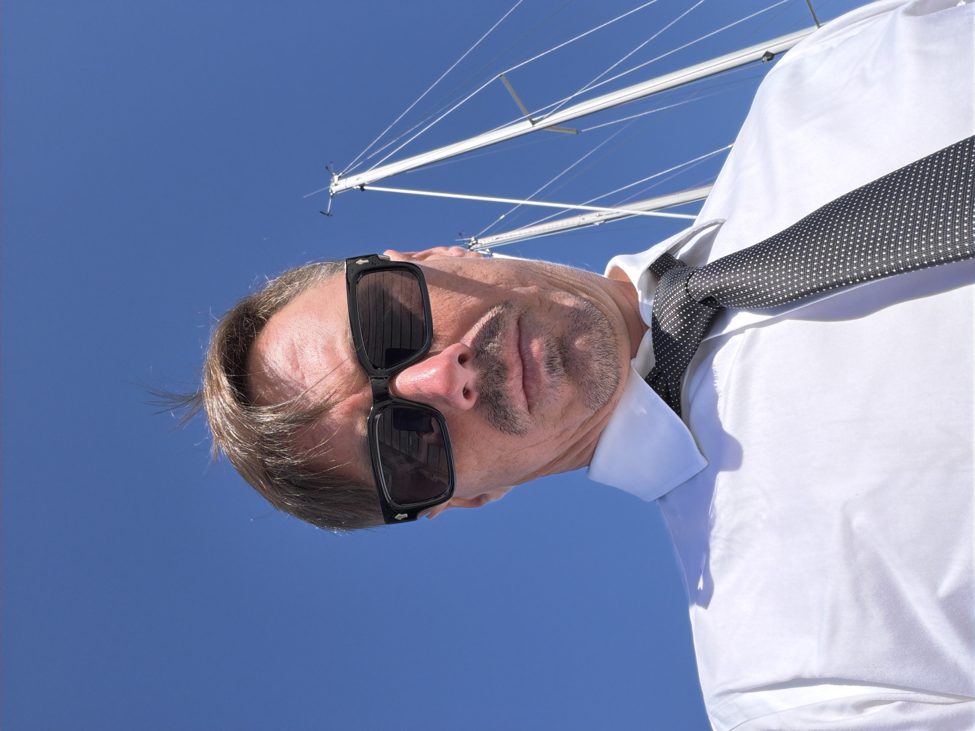 A low-angle, close-up selfie of a Caucasian man wearing a white collared shirt and a dark-patterned tie is captured against a clear blue sky. He has short brown hair, some of which is sticking up, and is wearing dark sunglasses. A faint mustache and goatee are visible. Behind him, the white masts and rigging of two sailboats are prominent against the sky. The man appears to be standing on a boat, as indicated by the visible rigging and masts. The lighting suggests it is a sunny day. The location is the Village of Sag Harbor, United States of America.