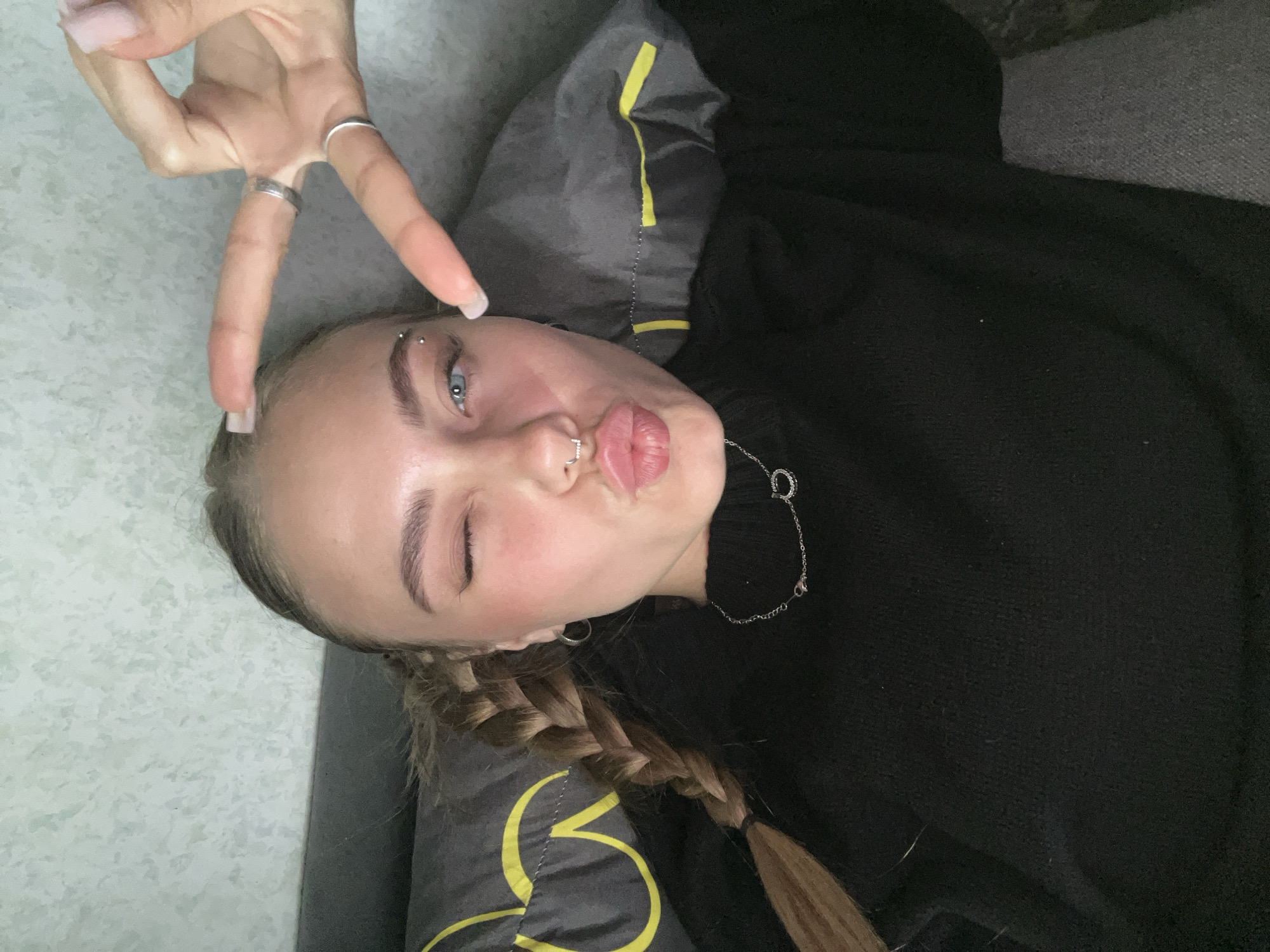 A young woman is depicted lying down, possibly on a couch or bed, in an indoor setting in Fokino, Russia. She is facing upwards towards the camera, with her head resting on a dark gray pillow featuring a yellow trim or abstract pattern. Her light brown hair is styled into two braids, one of which is visible over her right shoulder.
The woman has light blue eyes, and is captured in a playful pose: she is winking with her right eye and puckering her lips in a "kiss" gesture. Her left hand is raised, making a peace sign with two fingers. She wears a black long-sleeved top or sweater. Notable accessories include a barbell piercing with two studs above her right eyebrow, a septum ring with small gems in her nose, and a small hoop earring in her left ear. She also wears two silver rings on the fingers forming the peace sign and a silver chain necklace with a circular pendant around her neck. The background consists of a light gray, textured surface on the left and a darker, plain gray or black surface on the right. The overall mood of the image is casual and expressive. No visible text is present.