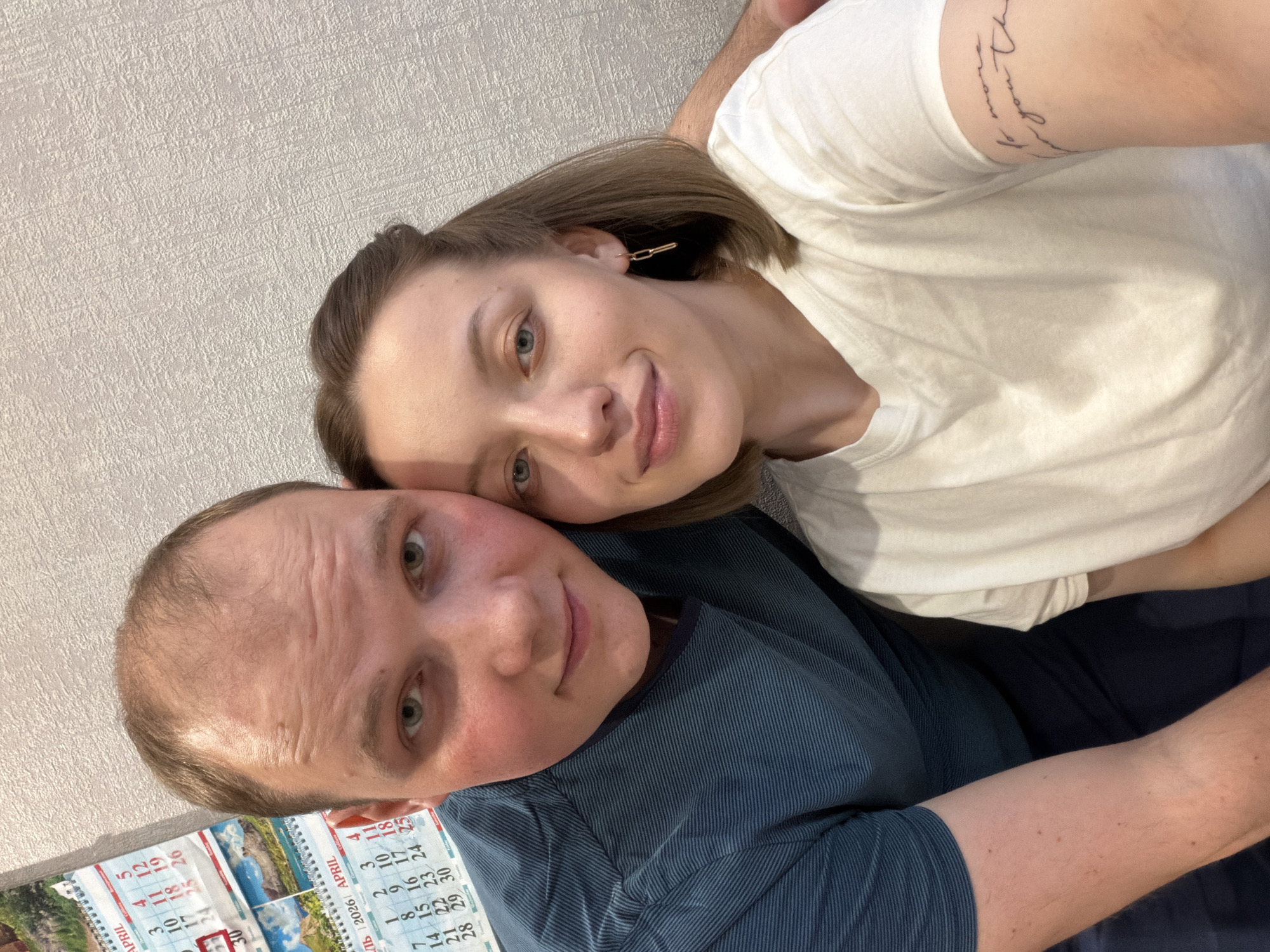 This media file presents a selfie-style portrait of a man and a woman, likely a couple, in a close and affectionate pose. The scene appears to be an indoor setting, identified by the user as Reshetnikovo, Russia.
The woman, positioned behind and to the right of the man, has light brown hair and is wearing a white t-shirt. She features a golden chain-link earring on her right ear and a script tattoo on her upper left arm, the text of which is illegible. She has light-colored eyes and a subtle, content smile, with her head gently resting on the man's shoulder.
The man, prominent in the foreground, has short brown hair with a receding hairline and is dressed in a dark blue shirt with fine vertical stripes. He also possesses light-colored eyes and a soft smile, looking directly at the viewer. Both individuals display emotions of happiness and warmth.
The background shows a light-colored wall covered with textured wallpaper. To the man's left, a spiral-bound wall calendar is partially visible. Visible text on the calendar includes "APRIL" and specifically "2026 APRIL", alongside various dates such as "1, 2, 3, 4, 5, 7, 8, 9, 11, 12, 14, 15, 16, 18, 19, 22, 23, 24, 25, 26, 29, 30", with some Cyrillic characters also present. The lighting suggests an indoor environment but does not indicate a specific time of day.