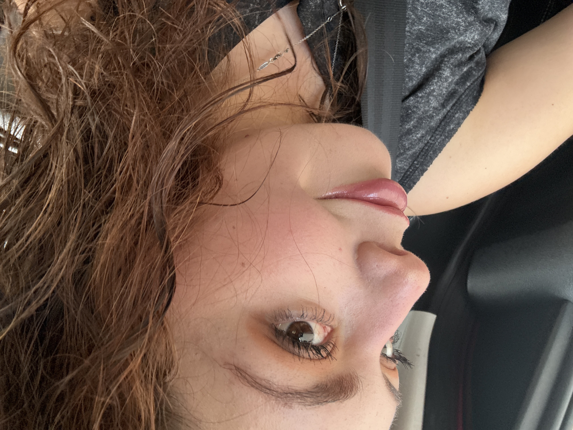 The image shows a close-up of a person's face, angled and rotated counter-clockwise. The person has brown hair that appears slightly damp. The skin is light-toned with visible pores. The makeup includes light eye shadow and glossy lips. A thin silver necklace is visible. The person is wearing a dark gray shirt, and a seatbelt is visible diagonally across the body. The background includes a car interior. There is no information to determine if this image has anything to do with Santa Clara Ocoyucan, Mexico.