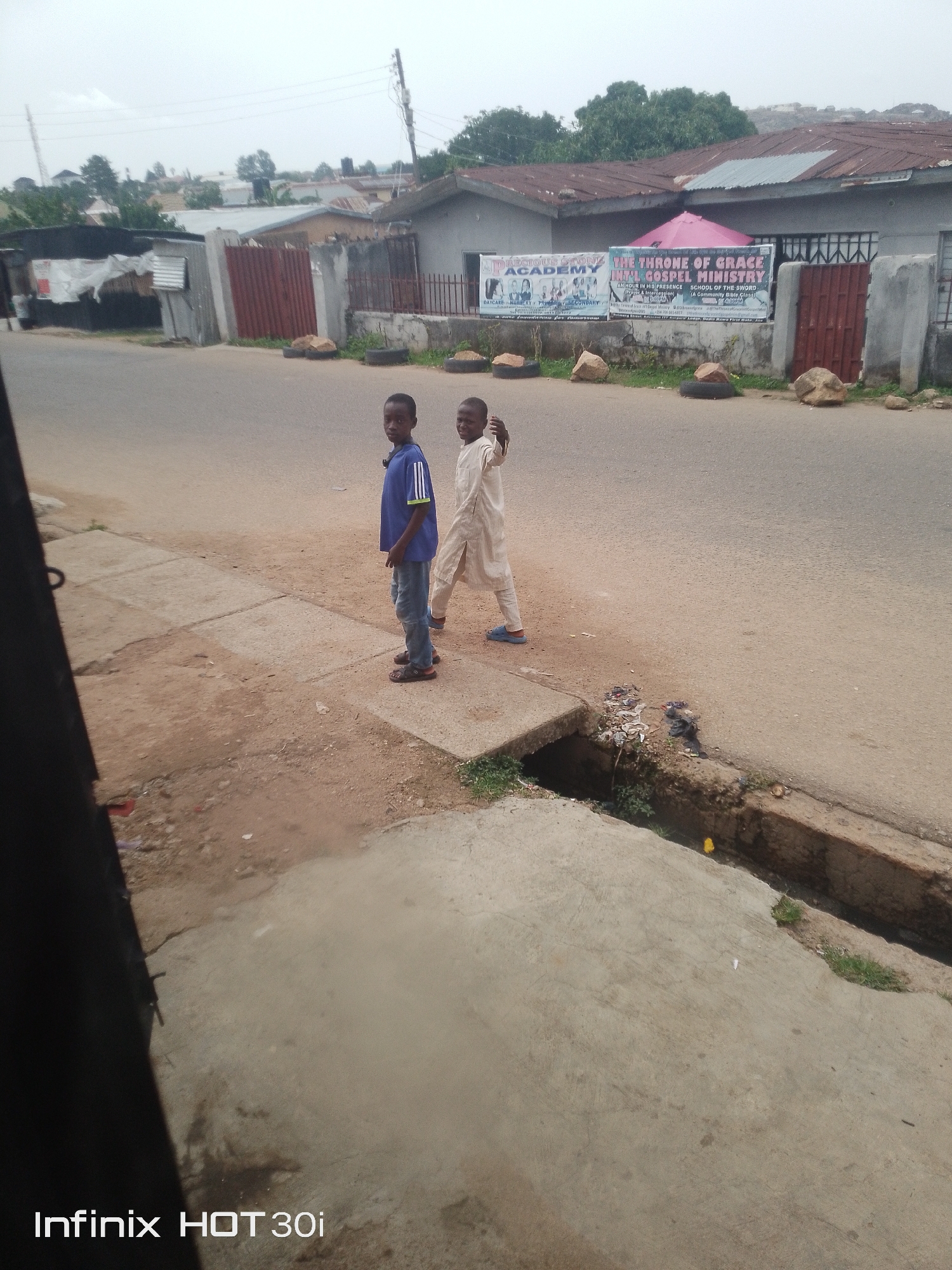 Two young boys are standing on a concrete sidewalk next to a paved road. The boy on the left is wearing a blue shirt with green stripes and blue jeans. The boy on the right is wearing a cream-colored outfit. Behind them, there is a building with a sign that reads "PRECIOUS STONE ACADEMY" and "THE THRONE OF GRACE". The area appears to be a residential or commercial street with houses and trees visible in the background. The sky is overcast, suggesting a cloudy day.