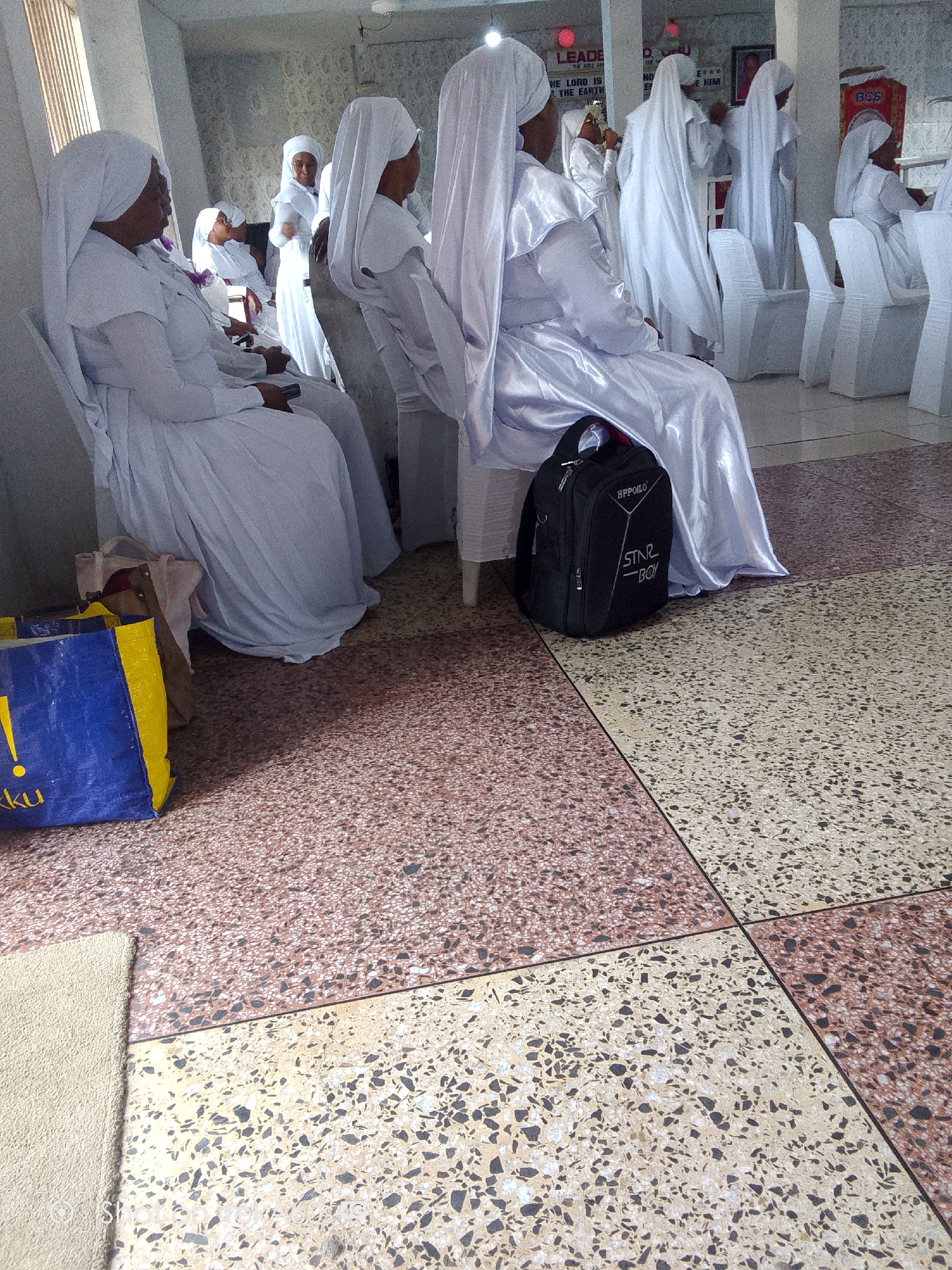 The image shows a group of women dressed in white robes and head coverings, likely attending a religious gathering. They are seated on white plastic chairs in an indoor space with a tiled floor. A black backpack with the inscription "HPPOILO STAR-BOY" sits next to one of the women. A blue and yellow shopping bag is on the floor near another woman. Based on the architectural style and the presence of the "BCS" logo (likely referring to the Brotherhood of the Cross and Star, a religious organization), the location is likely in Lagos, Nigeria. The atmosphere appears quiet and orderly.