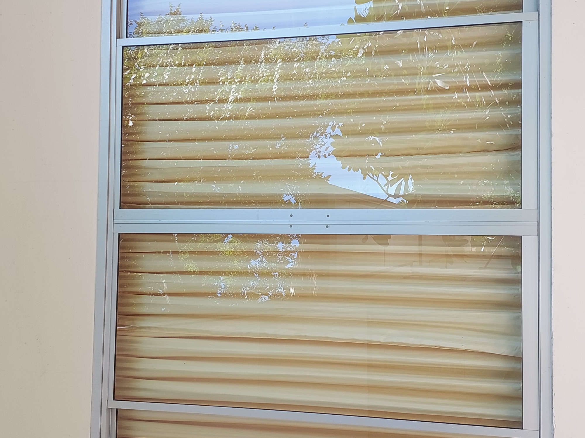 The image shows a sliding glass door with light yellow curtains partially drawn. The doors are framed by a silver aluminum structure. Reflections of tree branches and leaves are visible on the glass, indicating an outdoor environment. The background is a plain, light-colored wall. The location is identified as Baan Mae Nam, Thailand. There are no people or discernible actions in the image.