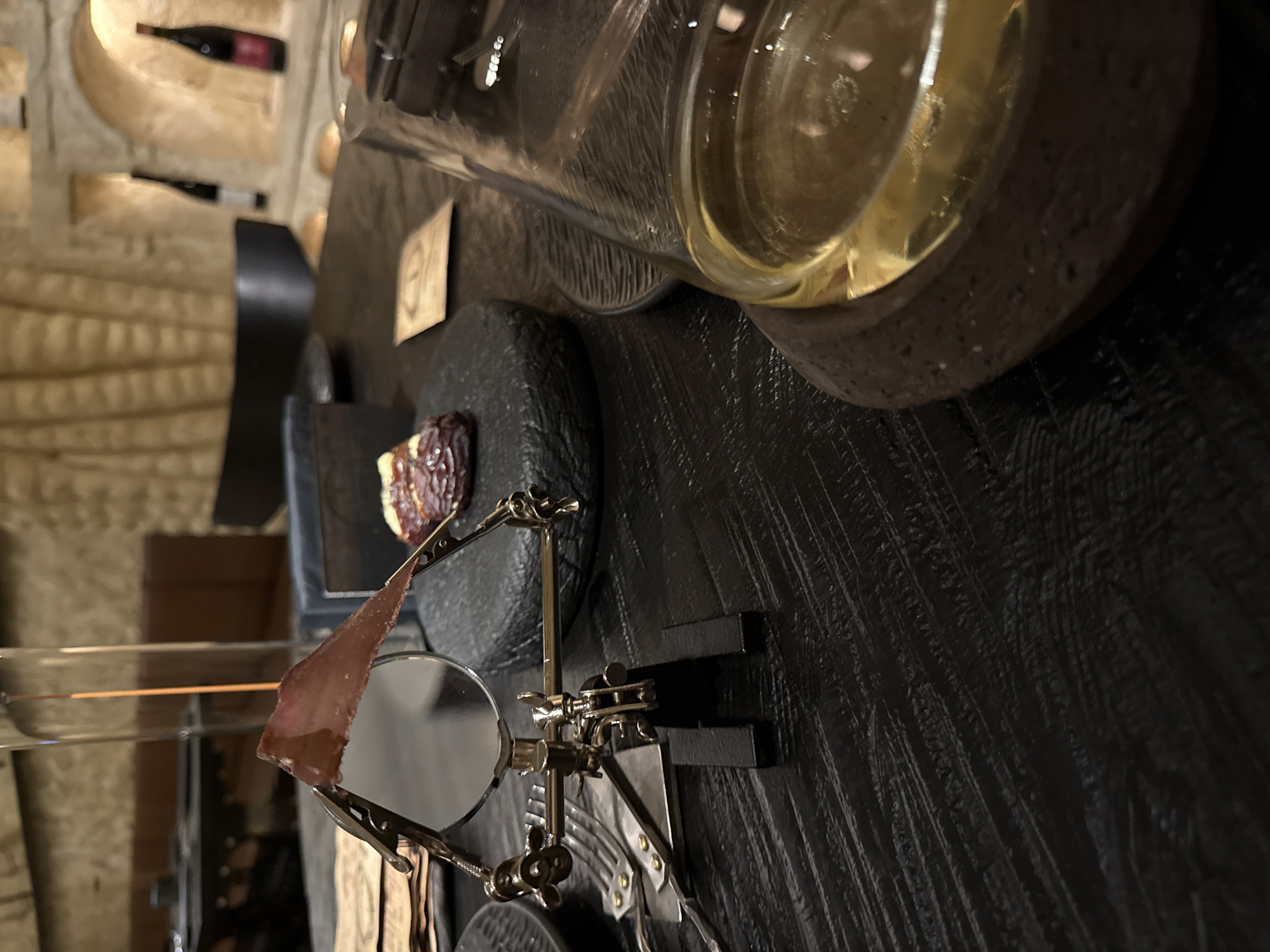 The image depicts a stylized food presentation, possibly in a restaurant setting. In the foreground, a thin slice of cured meat (likely prosciutto or a similar product) is held by a metallic stand with a small magnifying glass. Behind the meat, a small black stone plate presents a date or similar fruit. A large glass container filled with a yellowish liquid rests on a dark, textured surface, possibly a wooden table.
In the blurred background, there are indications of a stone-walled environment, with niches containing wine bottles. The overall composition suggests a sophisticated or artistic food experience.
While the prompt asks about the location, there is no definitive evidence within the image to confirm it is Moscow, Russia. Therefore, I cannot accurately state the location.