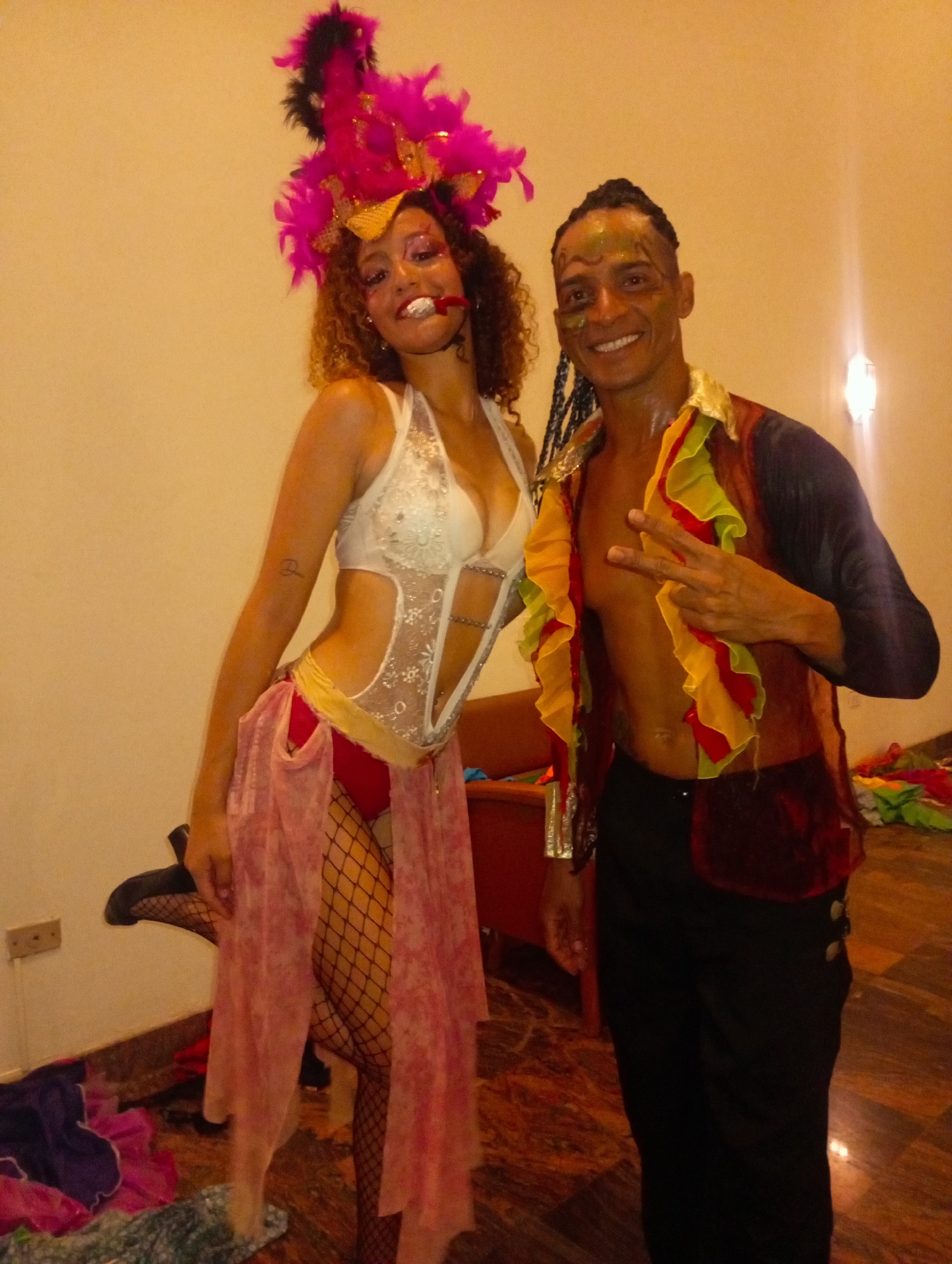 Two people are posing in a room. A woman on the left is wearing a white halter top, red bottoms, fishnet stockings, a sheer pink skirt, and a pink and black feather headdress. A man on the right is wearing black pants, a dark shirt, and a vest with yellow and red ruffles. He has braids and painted designs on his face. He is making a peace sign with his hand. The room has a light-colored wall and a patterned floor. Scattered clothing and a red bench are also visible in the background. No evidence suggests the location is Pedro González, Venezuela.