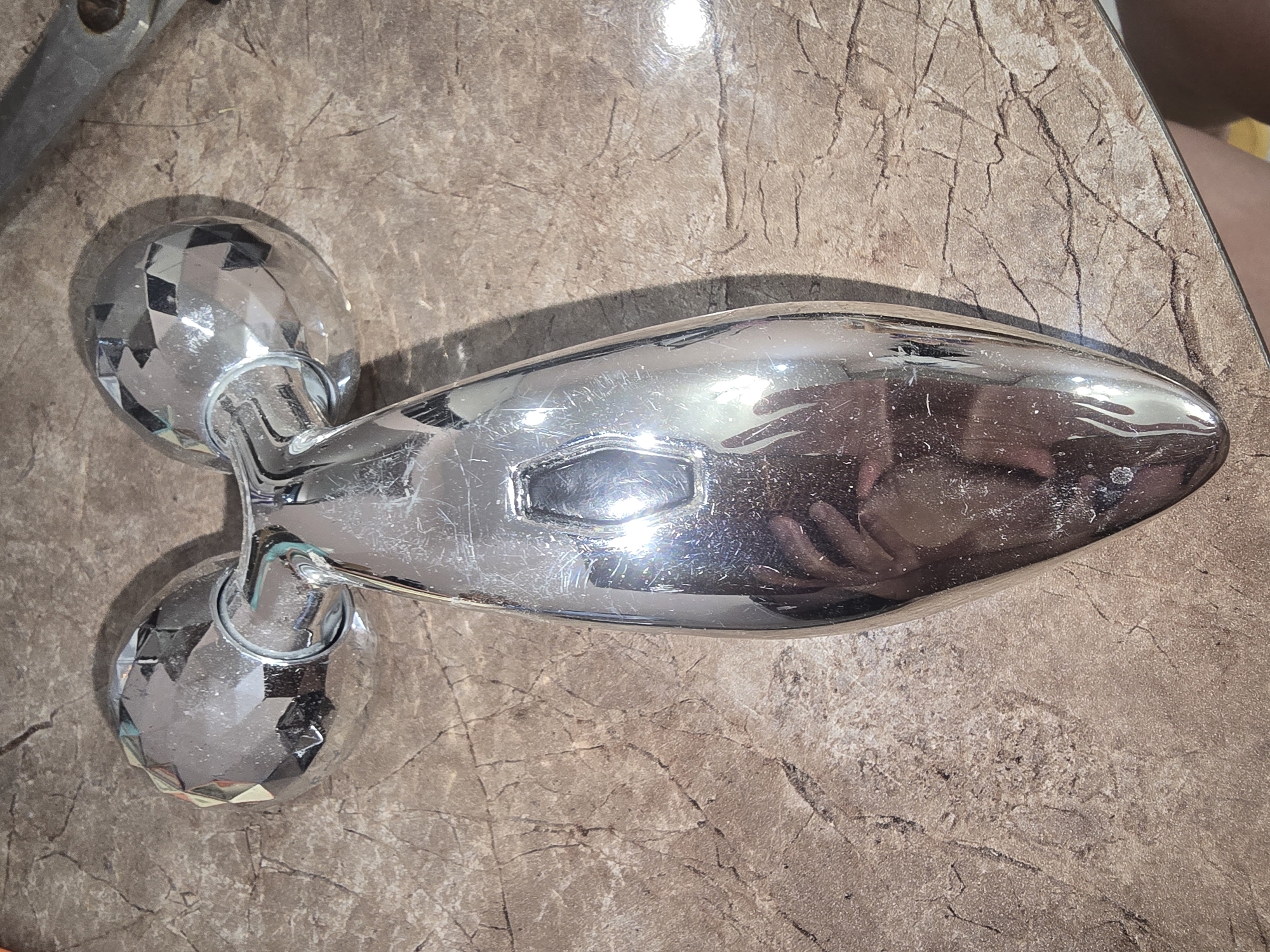 The image shows a close-up, overhead view of a silver facial roller tool resting on a marble-patterned surface. The tool has two faceted, spherical rollers at the top, connected by a metallic handle that tapers to a rounded point. The surface is a light brown color with darker brown and black veining, resembling cracked marble. The lighting in the image is bright, causing reflections on the metallic surface of the roller. The background is blurred, but part of a silver object with a dark handle is visible on the right side. The overall scene appears to be indoors, on a tabletop or similar flat surface. There are no indications of the location being in Ulan-Ude, Russia.