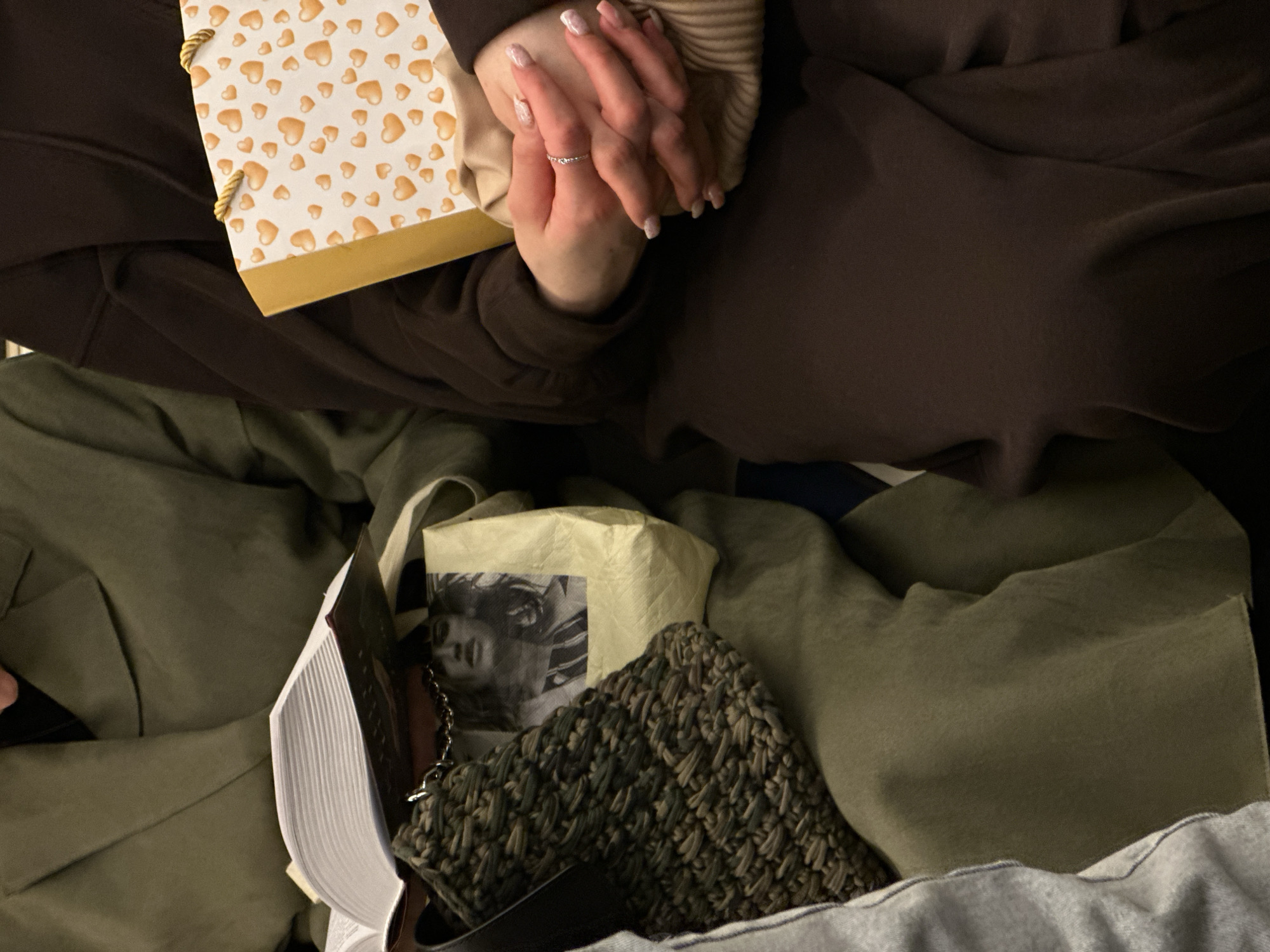 The image depicts a person seated with a brown outfit, holding a gift bag with a pattern of gold hearts. Beneath them is a collection of items including an open book, a tote bag with a black and white image, and a knitted bag. The location appears to be an indoor setting in an Unknown city.