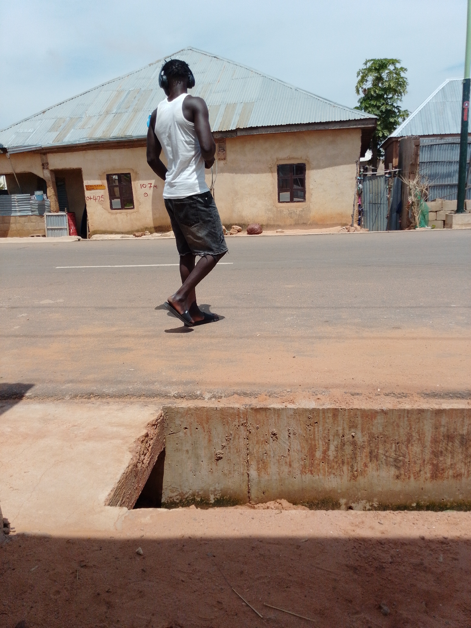 A man wearing a white tank top, dark denim shorts, and black flip-flops walks down a paved road. He has headphones on and is looking to the right. In the background, there are two single-story buildings with corrugated metal roofs. The building on the left has a light-colored exterior, a dark-framed window, and a sign with writing that is partially obscured. The building on the right has a similar style, with a corrugated metal fence. A large tree is visible between the two buildings. The foreground of the image shows a concrete drainage ditch filled with sand and some debris. The road surface is dusty and worn, with a white line marking the center. The sky is overcast.