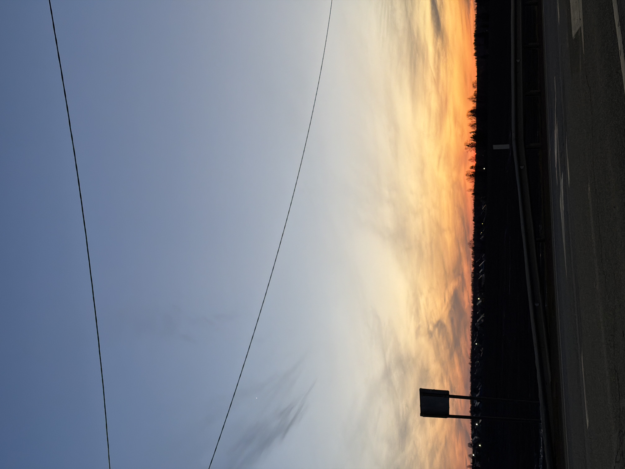 The image shows a sunset scene near a road in Kuregovo, Russia. Overhead power lines cross the pale blue and orange sky. A dark treeline and faint lights are visible in the distance, along with a road sign. The edge of a road runs along the right side of the frame.