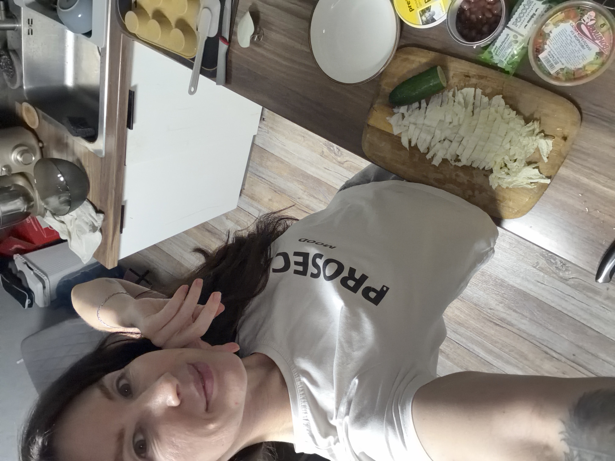 The image displays a female subject taking a selfie in an indoor kitchen environment. The woman, with dark hair and a light complexion, looks directly at the camera with a slight smile, holding her right hand near her face. She wears a light-colored t-shirt with the black text "PROSEC MOOD" on the chest. A silver-colored bracelet is visible on her right wrist, and a dark tattoo is partially visible on her left upper arm.
The scene focuses on a dark wood-grain kitchen countertop where food preparation is underway. A wooden cutting board holds a pile of finely chopped light-colored leafy vegetables, likely cabbage, next to a partially sliced green cucumber. To the right of the cutting board, a white round plate with a brown rim is present. Adjacent to these items are several food products: a yellow round container marked "premia" (ПРЕМИЯ), an open small plastic container with dark beans, a green packet labeled "СОУС НАТУРАЛЬНЫЙ" (Natural Sauce) and "РОССОЯ" (Rossiya), and a clear plastic container with "САЛАТ КОРЕЙСКИЙ ИЗ МОРКОВИ" (Korean Carrot Salad) and "Оволожка" (Ovolozhka) in Cyrillic script. Further right, a yellow silicone baking mold and two spatulas (one grey, one white) are visible.
In the background, a stainless steel kitchen sink is set into the countertop above white kitchen cabinets. A chrome stand mixer and a dark grey bowl are positioned to the left of the sink, and a light blue bowl is to its right. The flooring is light wood-look laminate. To the upper left, a portion of a grey upholstered couch or chair is visible. In the upper right background, a white and black athletic shoe rests on a surface, next to a red object. The specific location within the city of Perm, Russia, cannot be determined from the visual evidence presented in the image.
