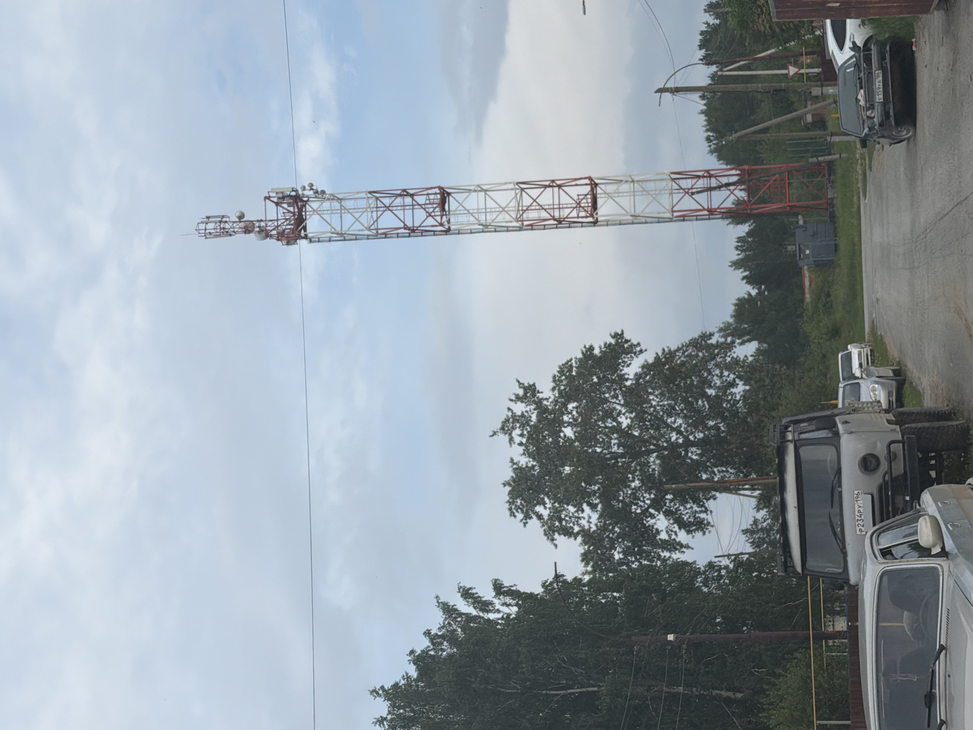 A communication tower stands tall against a cloudy sky in a rural or suburban area. The tower is red and white, with various antennas and equipment attached to its structure. In the foreground, several vehicles are parked along a dirt road. These include a light-colored sedan in the left, a gray utility vehicle with large tires behind it, and a black car with its hood open on the right. Trees and greenery surround the area, with a fence visible on the left side of the road. The scene appears to be in a semi-developed area, possibly on the outskirts of a town or village.