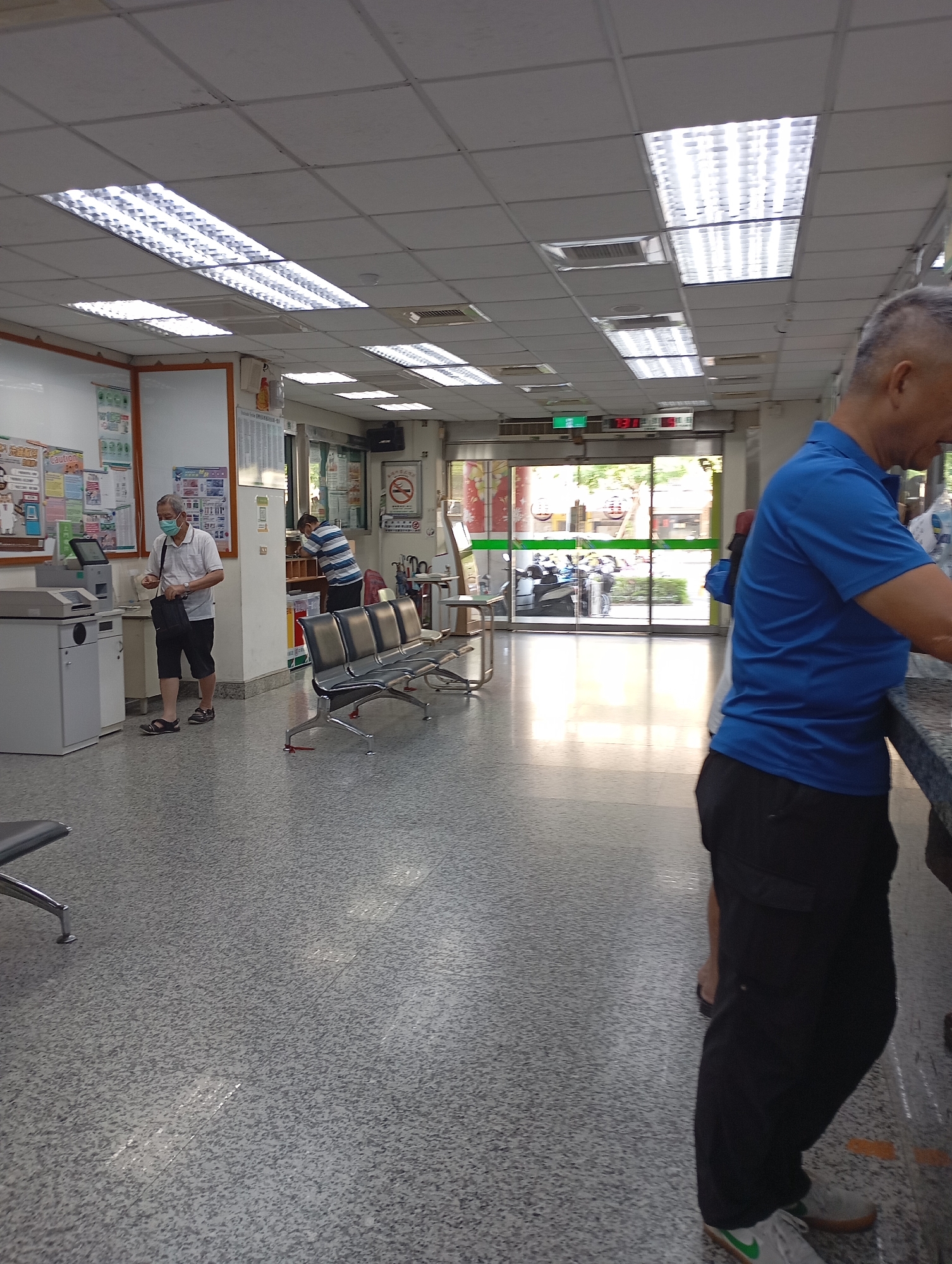 The image depicts the interior of what appears to be a clinic or hospital waiting area in Kaohsiung, Taiwan. Several individuals are present: one man wearing a mask and carrying a black bag is interacting with a payment kiosk. Another man in a striped shirt is standing by a counter. A man in a blue shirt and black pants is standing at a counter near the camera. There are rows of metal benches for seating. The room is well-lit by fluorescent ceiling lights. Signage is visible in Traditional Chinese. The exterior is visible through a glass entrance; motorbikes are parked outside.