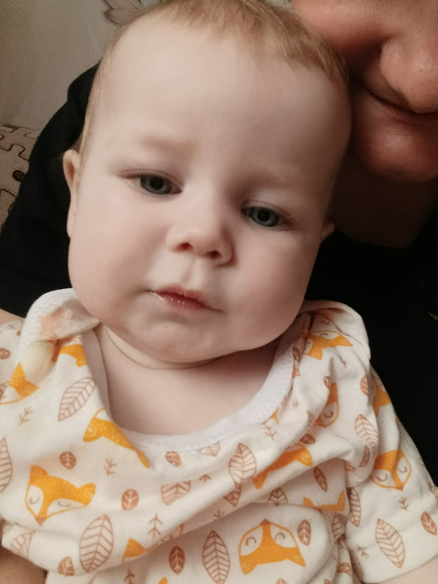 A close-up shot features a baby with fair skin, light-colored hair, and blue eyes. The baby is wearing a light-colored shirt patterned with images of foxes and leaves. A portion of an adult's face is visible in the upper right corner of the frame. The background appears to be a dark, neutral color, with a glimpse of patterned fabric behind the baby's left shoulder. The photograph is taken at a high angle, focusing on the baby's face and upper body. The city of Unknown, Unknown is not discernable.