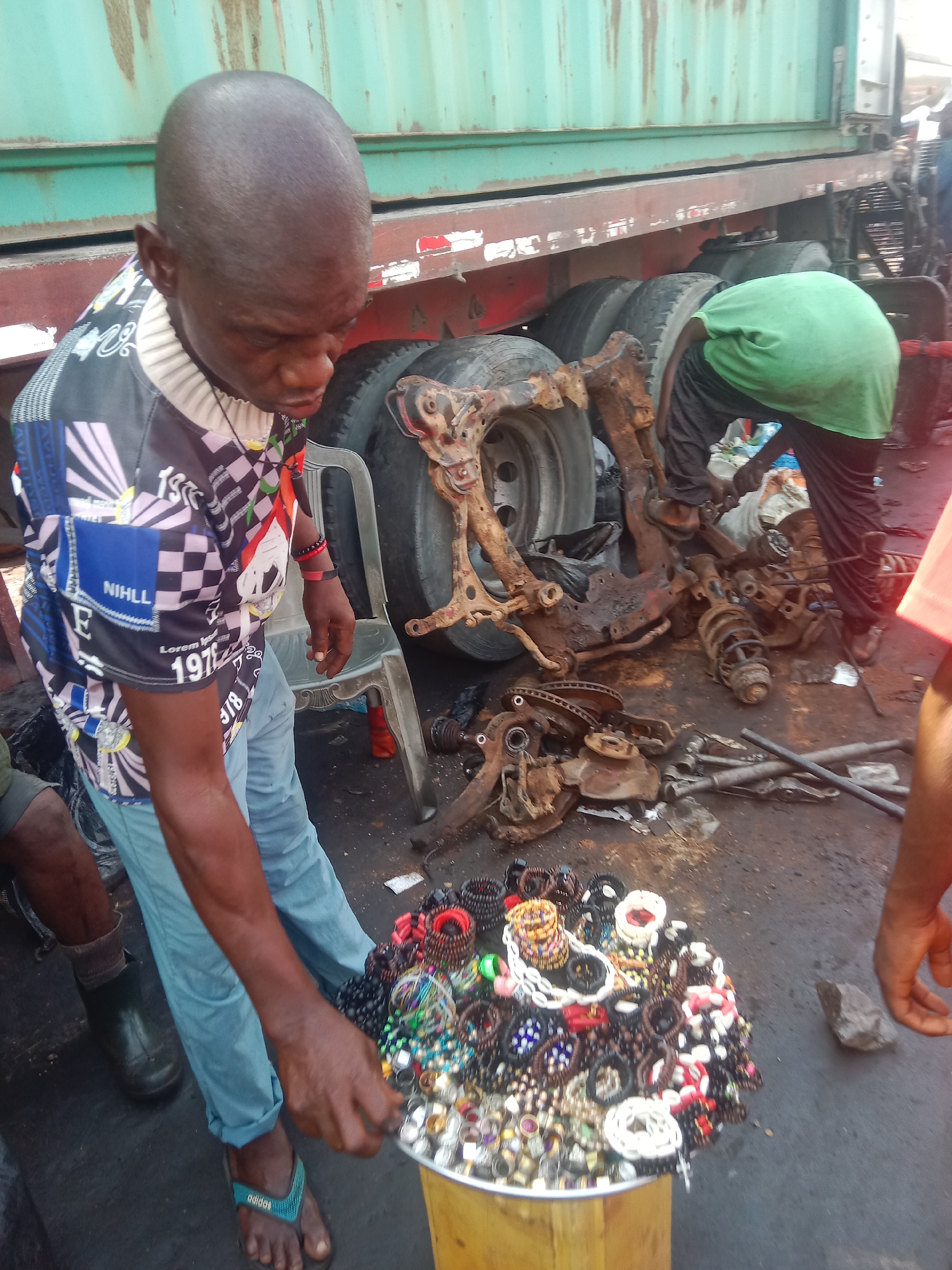 The image depicts an outdoor scene, likely a bustling roadside or informal repair area in Onitsha, Nigeria, under bright daylight. In the foreground, a bald man with dark skin, wearing a multi-colored patterned long-sleeve shirt, light blue pants, and teal Adidas flip-flops, holds a round, silver-colored tray overflowing with a diverse assortment of colorful bracelets, rings, and other small jewelry pieces. He appears focused on the merchandise. Text visible on his shirt includes "NIHL L" and "Lorem 197".
Behind him, a second man in a bright green t-shirt and dark pants is bent over, actively engaged in working on the undercarriage of a large, weathered green shipping container mounted on a red truck chassis. The ground is dirty and scattered with numerous rusted and disassembled heavy-duty vehicle components, including axles, tires, suspension springs, shock absorbers, brake rotors, and various chassis parts, suggesting a mechanical repair or salvage operation. A plastic chair and some tools are also visible amidst the debris. The scene conveys a sense of industrious activity in an urban or semi-urban setting.