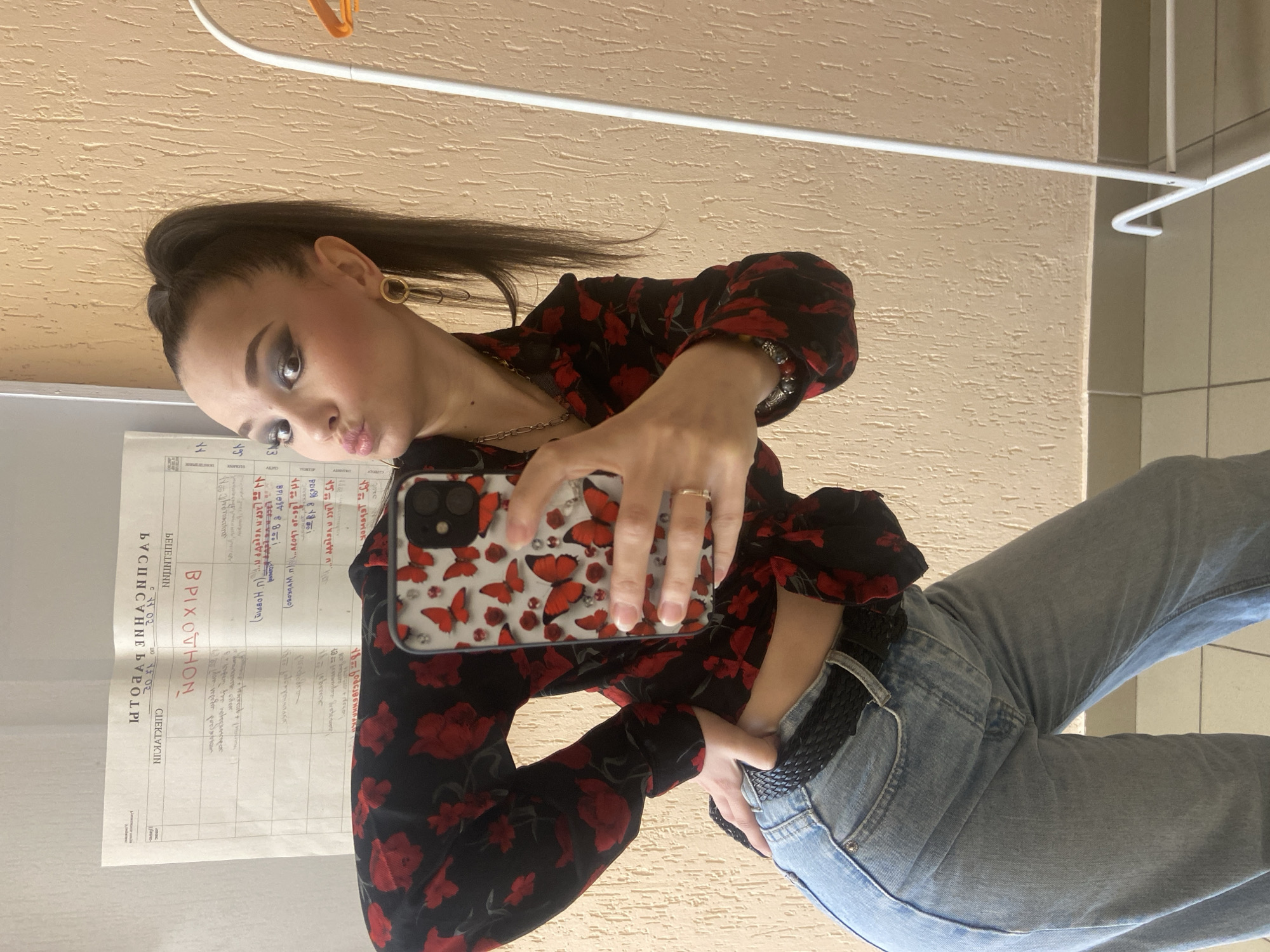 A young woman is taking a selfie indoors. She is wearing a black top with red flowers, light blue jeans, and a black belt. Her hair is pulled back in a ponytail and she is wearing earrings. She is holding a phone with a case that has red butterflies on it. There is a document in the background with Cyrillic text on it. The text "ВРИХОТНОН" is highlighted in red. The document appears to be a registration or a sign for a business named "ВРИХОТНОН." The city of Chaikovsky, Russia is not discernible from this image.
