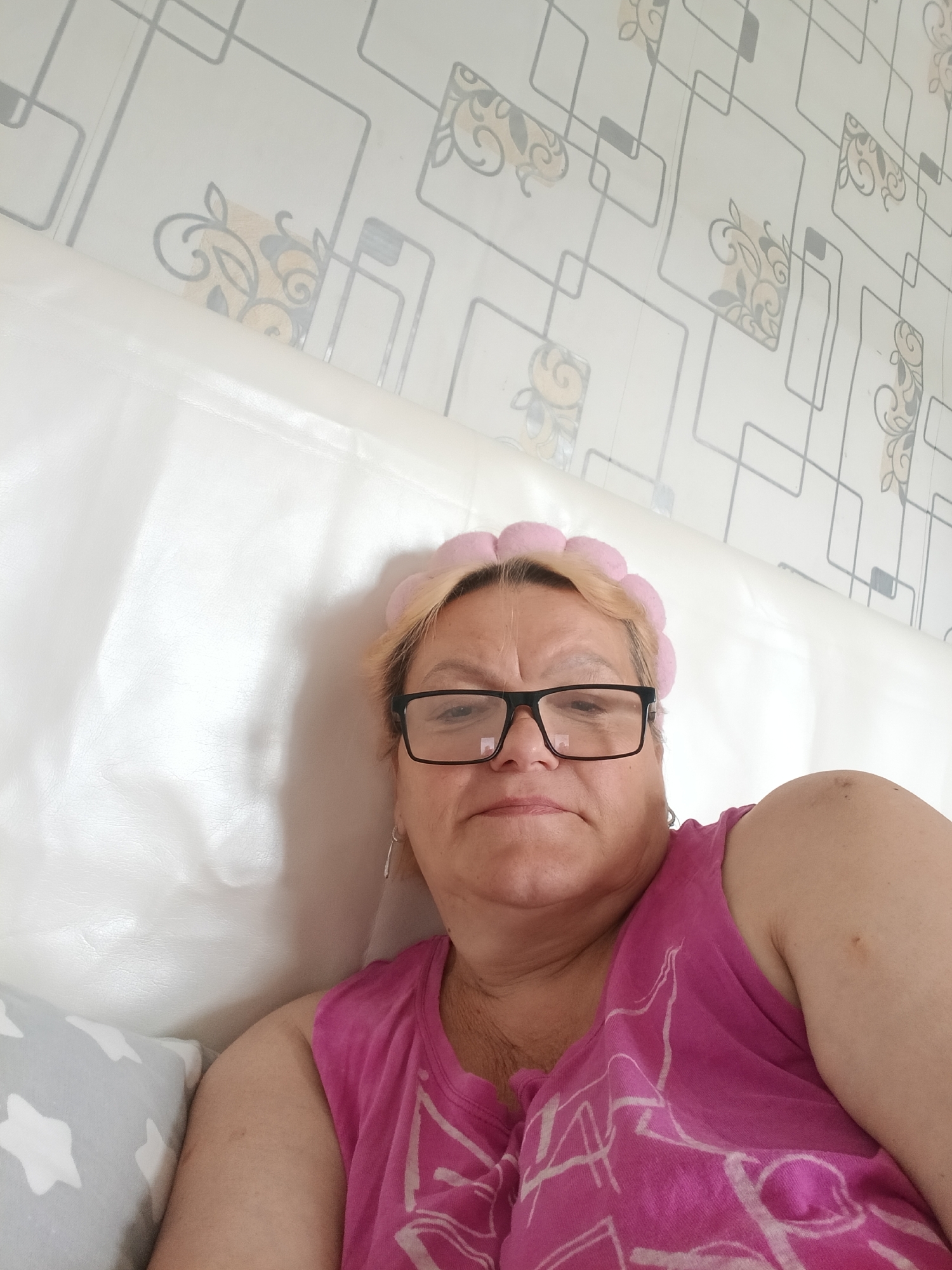The image shows a woman wearing glasses, a pink headband, and a pink shirt with a white pattern. She is lying against a white headboard or cushion, with a patterned wallpaper in the background. A pillow with white stars on a gray background is visible in the lower left corner. The location is indicated as Красный Маяк, Russia, although the image itself provides no direct visual confirmation of this.