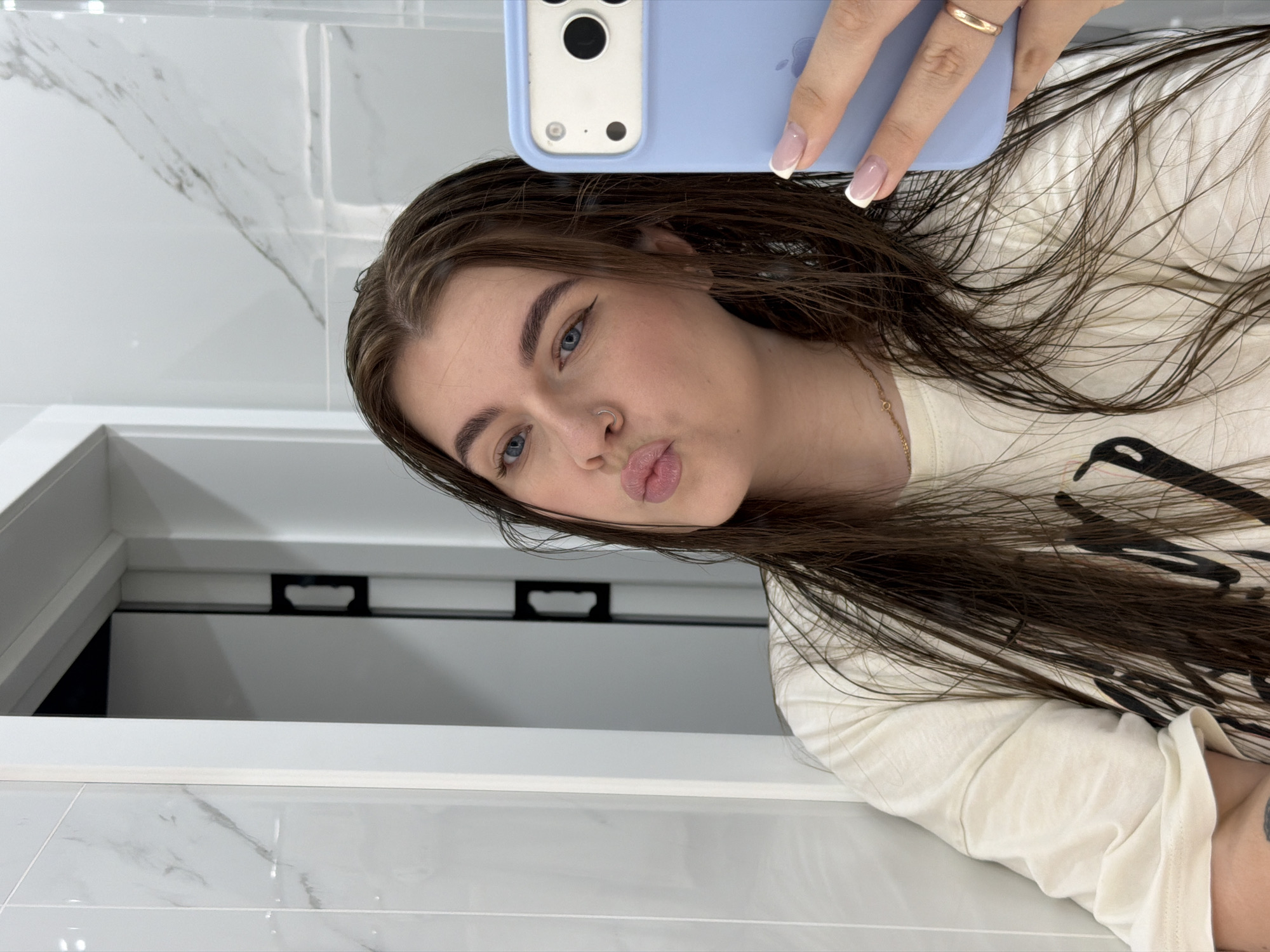 A young woman with long, dark brown hair and blue eyes takes a mirror selfie in a bathroom. She is wearing a light-colored t-shirt and has a nose ring and winged eyeliner. Her lips are pursed, and she is holding a light blue phone. The bathroom has marble-patterned tiles on the wall and a white door. The image appears to be taken in Chaikovsky, Russia, as indicated by the user's request.