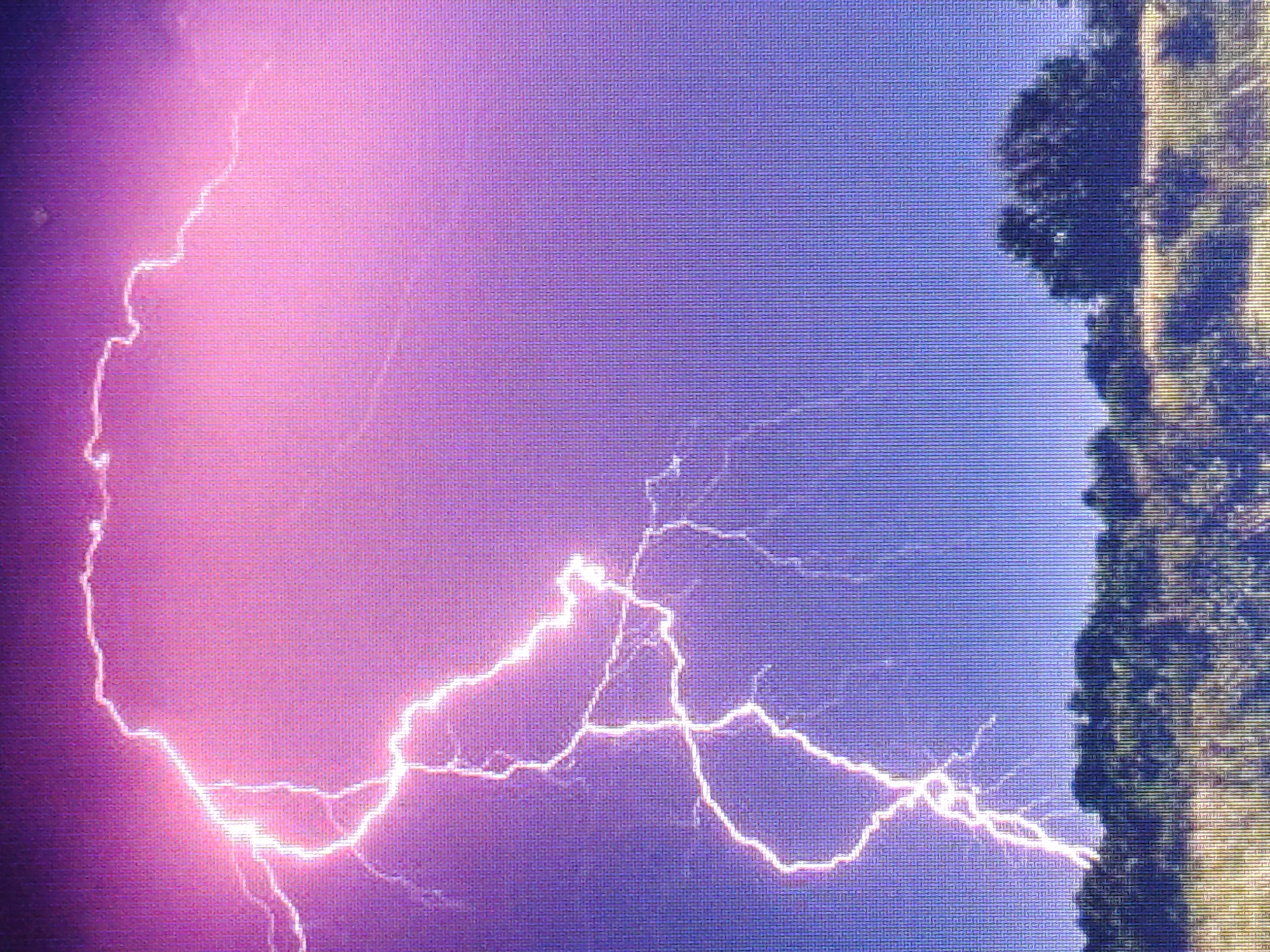 A dramatic lightning strike illuminates a dark, stormy sky with vibrant pink and purple hues. The jagged bolt of electricity branches out, a stark contrast against the ominous clouds. Below, a silhouetted landscape of trees and rolling hills suggests a rural or undeveloped area. The image captures the raw power and beauty of nature during a thunderstorm.