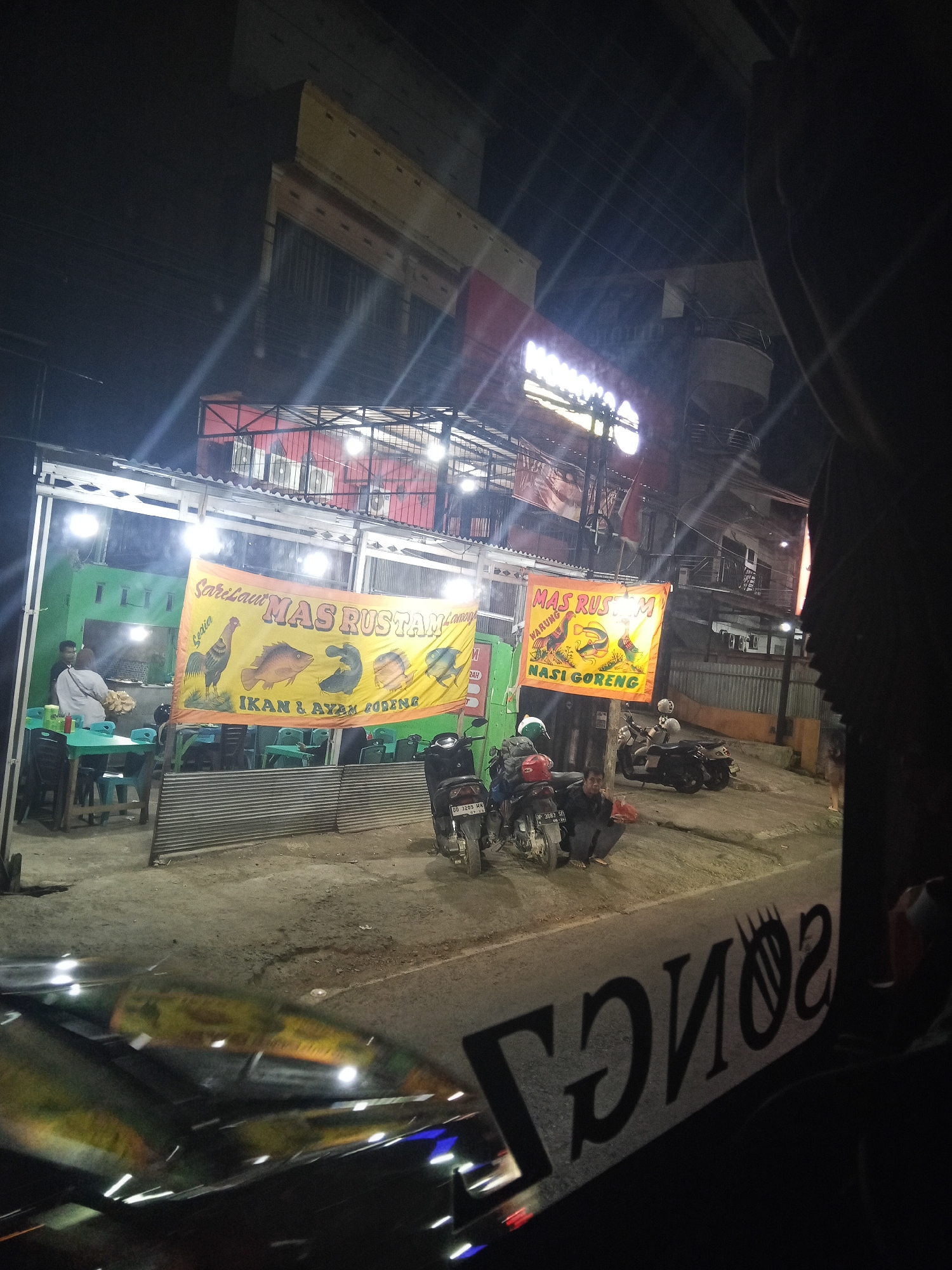 The image depicts a street scene at night in what appears to be a commercial area, possibly in Hasanuddin, Indonesia, as indicated by the text on a banner. In the foreground, a portion of a vehicle's windshield is visible, reflecting lights and the street ahead, with a dark interior framing the view.
Dominating the mid-ground are two banners. The one on the left is yellow and features a rooster and various fish illustrations, with text that reads "MAS RUSTAM" and "Sari Laut Ikan & Ayam Goreng" (Seafood, Fried Fish & Chicken). This banner is hung in front of a green building with a corrugated metal roof. Several tables and chairs are set up on a concrete area in front of this building, and two individuals are present, one sitting at a table and another standing.
To the right of the first banner is another yellow banner, this one shorter, with text that includes "MAS RUSTAM" and "NASI GORENG" (Fried Rice). This banner is also hung in front of a building. Several motorcycles are parked in front of the buildings. One motorcycle in the foreground has a license plate that reads "DD 3283 MM". A person is seen crouching near the motorcycles.
The background shows multi-story buildings, suggesting an urban or semi-urban setting. The lighting is primarily from artificial sources, including streetlights and the illuminated signs. The overall impression is of a modest roadside eatery operating at night.