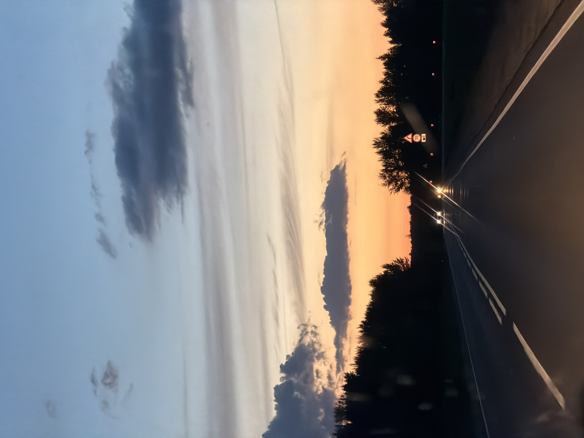 The image depicts a sunset scene on a road leading towards a forest. The sky transitions from a pale blue at the top to a warm orange hue near the horizon, with scattered clouds adding texture. A dark silhouette of trees lines the right side of the road, and on the left, the trees are less dense, allowing a glimpse of the sky. Two cars are visible on the road, with their headlights illuminated, suggesting it is dusk or early evening. A traffic sign indicating a speed limit of 70 is visible on the right side of the road, along with several orange road markers. The road itself is asphalt, with double white lines marking the lanes. The image is taken from a vehicle, as evidenced by what appear to be raindrops or smudges on the windshield in the foreground. The overall impression is one of a quiet drive through a rural landscape as the day ends. There are no people visible in the image. The location is identified as being in the city of Laishevo, Russia.