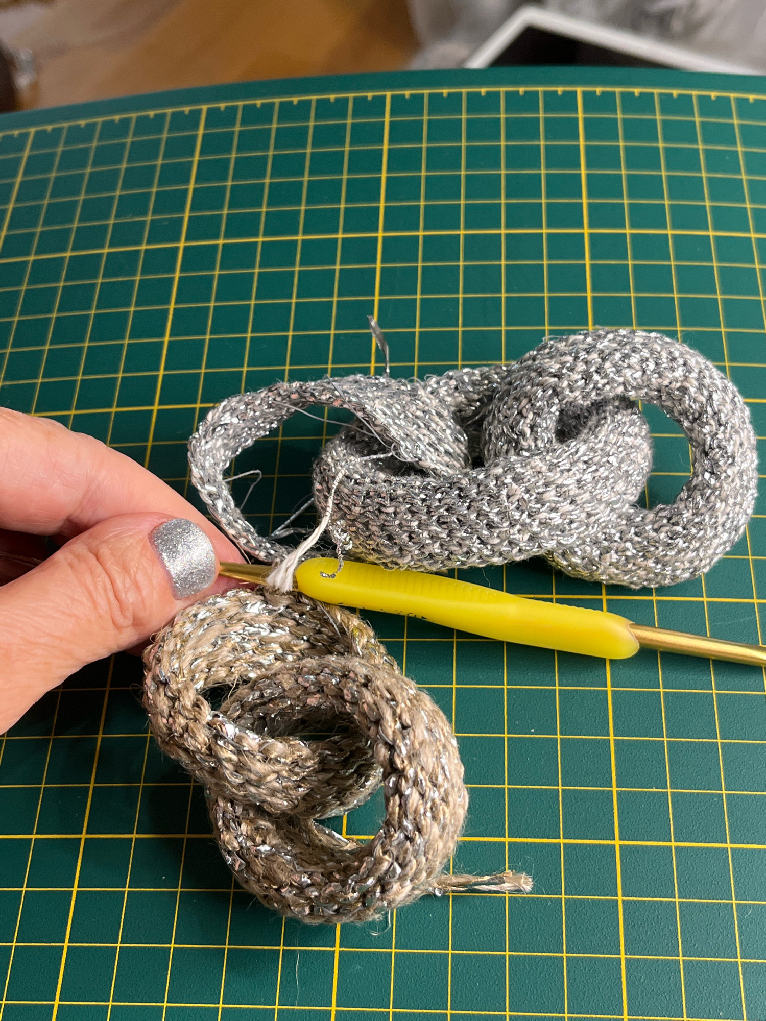 A close-up shot shows a person's hand with a sparkly silver fingernail holding a yellow crochet hook, which is partially inserted into a loop of brown and silver yarn. Adjacent to this, a finished crochet piece made of silver yarn forms a knotted shape. The objects are placed on a green cutting mat with a yellow grid pattern.