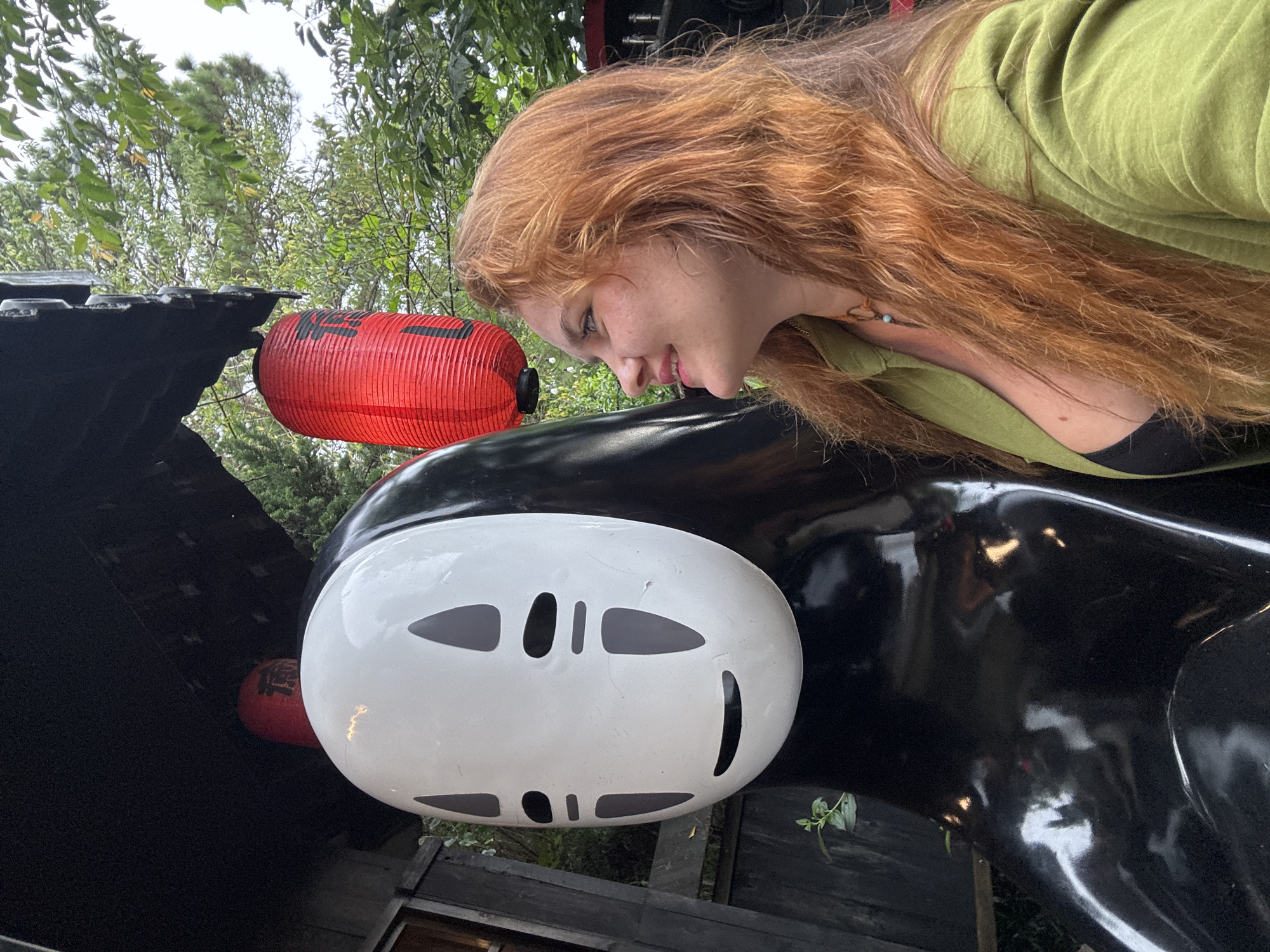 An outdoor medium shot shows a young woman with long, reddish-brown hair and fair skin, wearing a green jacket and a black top, standing next to a life-sized black statue of the character No-Face from Spirited Away. No-Face's iconic white mask with dark eyes and mouth is prominent in the foreground. A red lantern hangs above the statue. The background consists of lush green trees and foliage, suggesting a natural setting. The scene appears to be in the city of Cam Ly - Đà Lạt, Vietnam, a location known for its pine forests and natural beauty, potentially a park or a themed attraction.