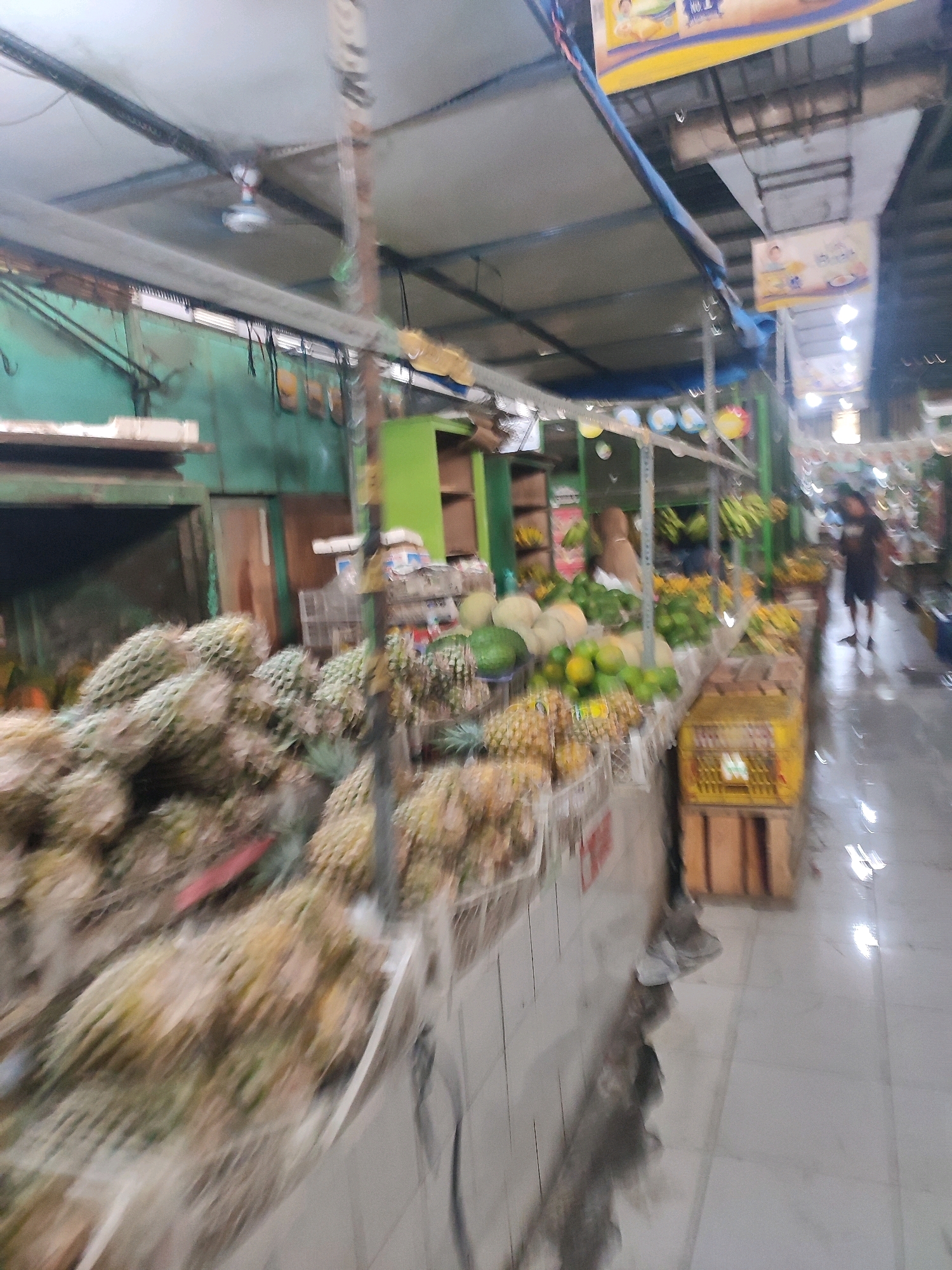 The image depicts a bustling market scene, likely located in Yogyakarta, Indonesia, given the prominent display of pineapples and other tropical fruits. The market appears to be an outdoor or semi-outdoor establishment with a covered ceiling supported by metal beams. A ceiling fan is visible, suggesting a humid climate. Stalls are laden with a variety of fruits, including a large quantity of pineapples in the foreground, as well as melons, limes, and bunches of bananas further down the aisle. Decorative banners with text are hung overhead. A person, seen from behind, is walking through the market. The floor is tiled and appears wet in some areas, reflecting the overhead lights. The overall impression is one of a vibrant, everyday marketplace.