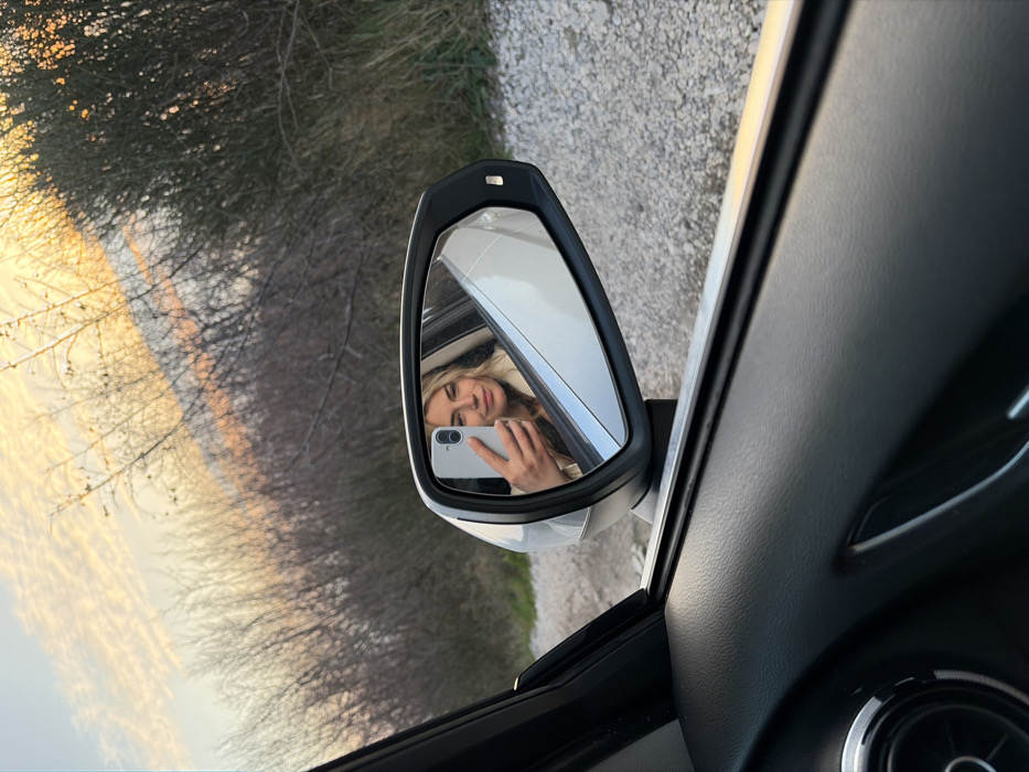 The image depicts a blonde female capturing a selfie reflected in the driver's side exterior mirror of a vehicle, positioned in Vrba, Montenegro. The perspective is from inside the car, looking out. The lower left of the frame shows the dark, textured interior panel of the car door. The white exterior mirror reflects the woman, who holds a light-colored smartphone in her left hand, aimed at the mirror. She has light-colored hair, a faint smile, and wears a light-toned garment. Beyond the vehicle, a gravel road or shoulder is visible, leading to a dense line of bare or sparsely leaved bushes and small trees. The sky above displays warm hues of orange and pink, characteristic of either sunrise or sunset.