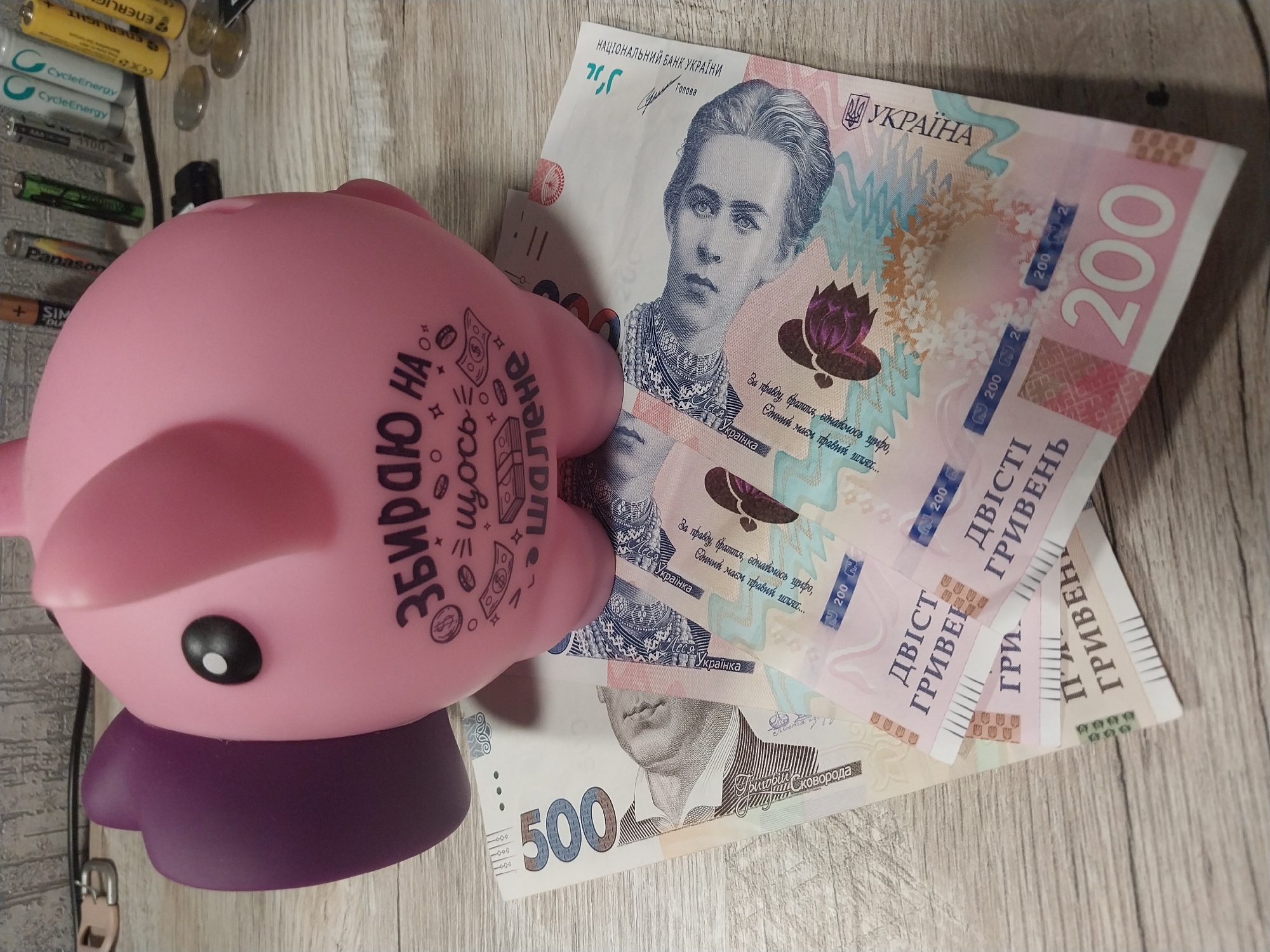 This is a close-up, high-angle shot of a pink ceramic piggy bank shaped like an elephant. The piggy bank is adorned with black text that reads "Збираю на щось шалене" which translates to "Saving for something crazy." Below the piggy bank are several Ukrainian hryvnia banknotes. Specifically, there is one 500 hryvnia note, and at least three 200 hryvnia notes. The banknotes are fanned out and partially obscured by the piggy bank. The background consists of a wooden surface with a light gray wood grain. To the upper right of the piggy bank, a collection of batteries and coins are visible. The overall scene is indoors, likely on a table or desk. There is no discernible location within the city of Dnipro, Ukraine, and no people are present in the image.