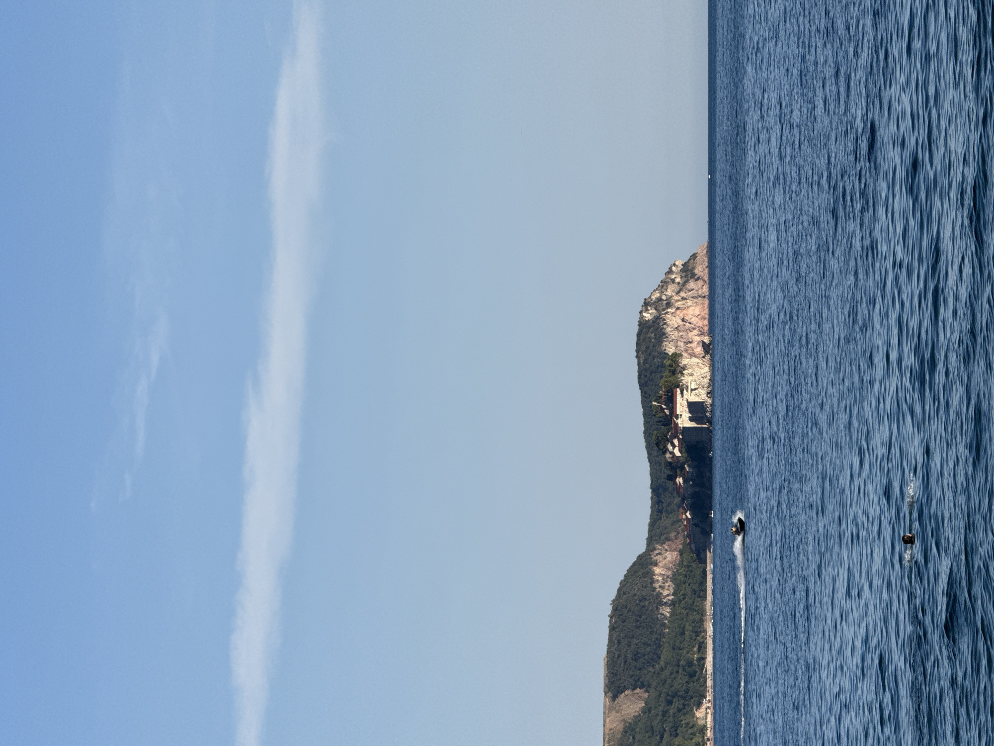 A wide, vertical shot captures a serene seascape with a coastal town nestled on a rocky promontory. The sky above is a clear, pale blue with a thin, wispy cloud stretching horizontally. The sea is a deep, rippling blue, with a person swimming in the foreground and a jet ski leaving a white spray in its wake further out. The coastal town, appearing to be in Rafailovići, Montenegro, is situated on a cliff, with buildings visible amongst lush green trees. The promontory itself is characterized by exposed reddish-brown rock faces interspersed with vegetation. The overall impression is one of a tranquil coastal scene on a sunny day.