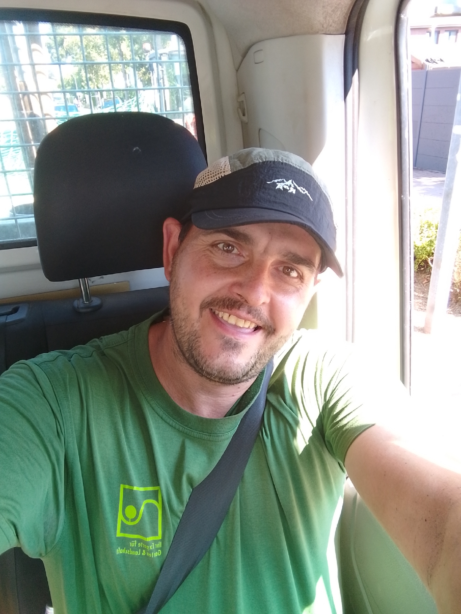 A man wearing a green t-shirt with a logo and a black and gray baseball cap is seated in a vehicle, taking a selfie. The t-shirt has a lime green logo with abstract shapes and text below it. The man has brown eyes, a mustache, and stubble. He is smiling, showing his teeth, and looking directly at the camera. A dark gray seatbelt is visible across his chest. The background shows the interior of the vehicle, including a headrest and a window with a protective grate. Through the window, trees and a building can be seen. The lighting suggests it is daytime, with sunlight entering the vehicle from the right side. The location is identified as Schenefeld, Germany, suggesting this is likely a residential or commercial area within the city.