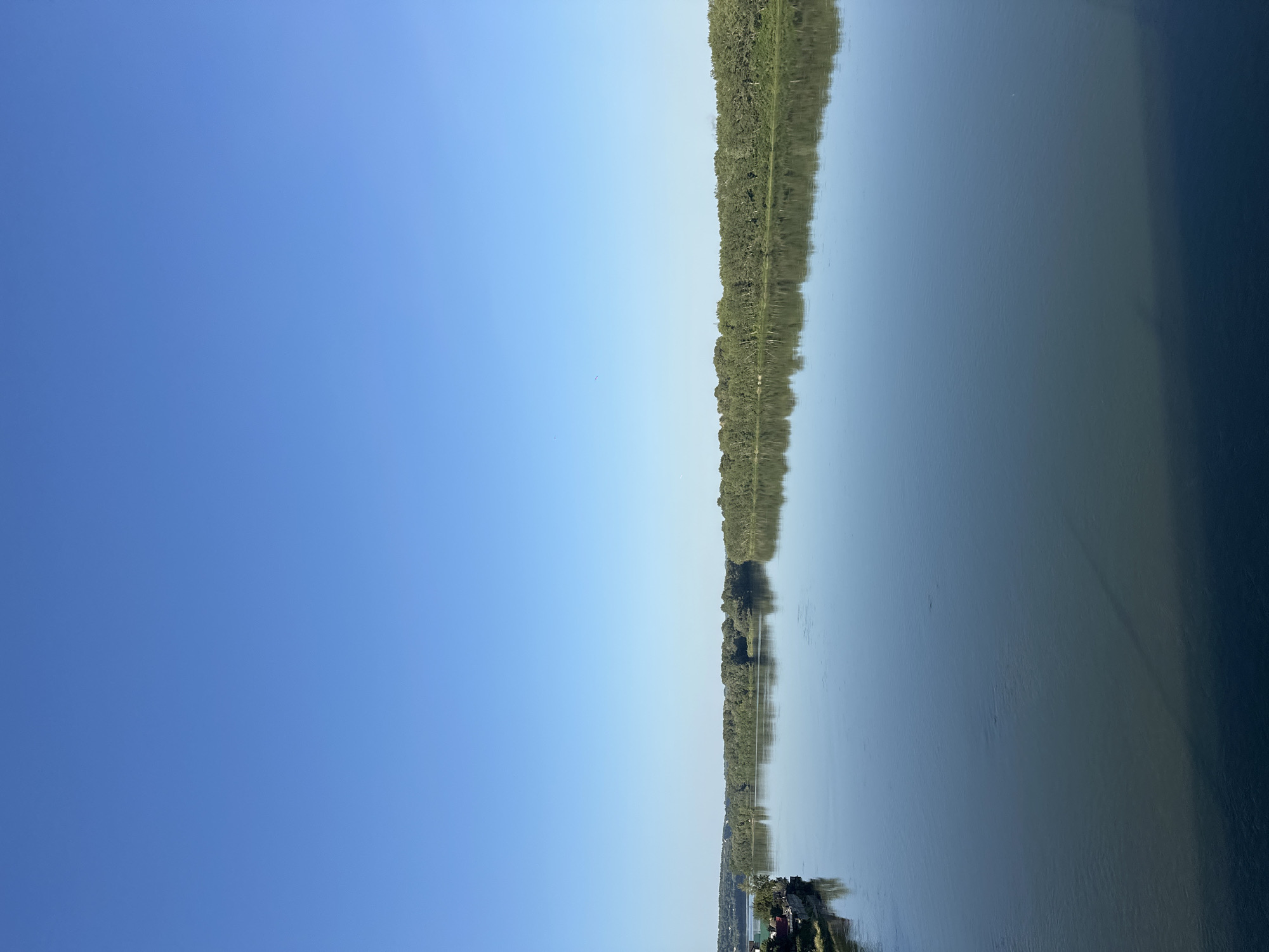 The image depicts a wide, calm river under a clear blue sky. The left bank of the river features a cluster of houses with red roofs, a small docked boat, and a line of trees. The opposite bank is densely lined with a thick forest of green trees, whose reflection is mirrored in the still water. The river itself occupies the majority of the frame, with its surface showing subtle ripples and variations in color, ranging from a deep blue in the foreground to a lighter, more reflective hue near the horizon. The overall impression is one of tranquility and natural beauty. This scene is located in the city of Biysk, Russia, on the Biya River.